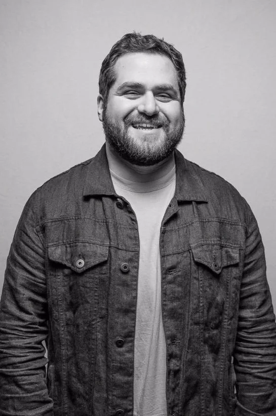 Black and white photo of a smiling man with a beard, wearing a denim jacket over a t-shirt, standing against a plain background.