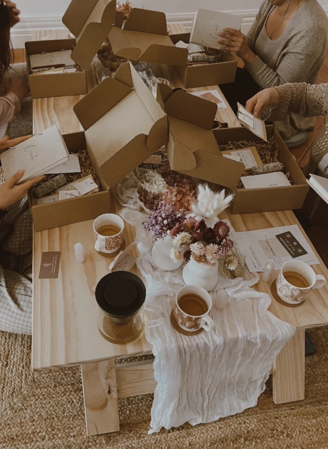People unpacking ritual hampers on a wooden table decorated with flowers, cups of tea, and papers, in a cozy indoor setting.