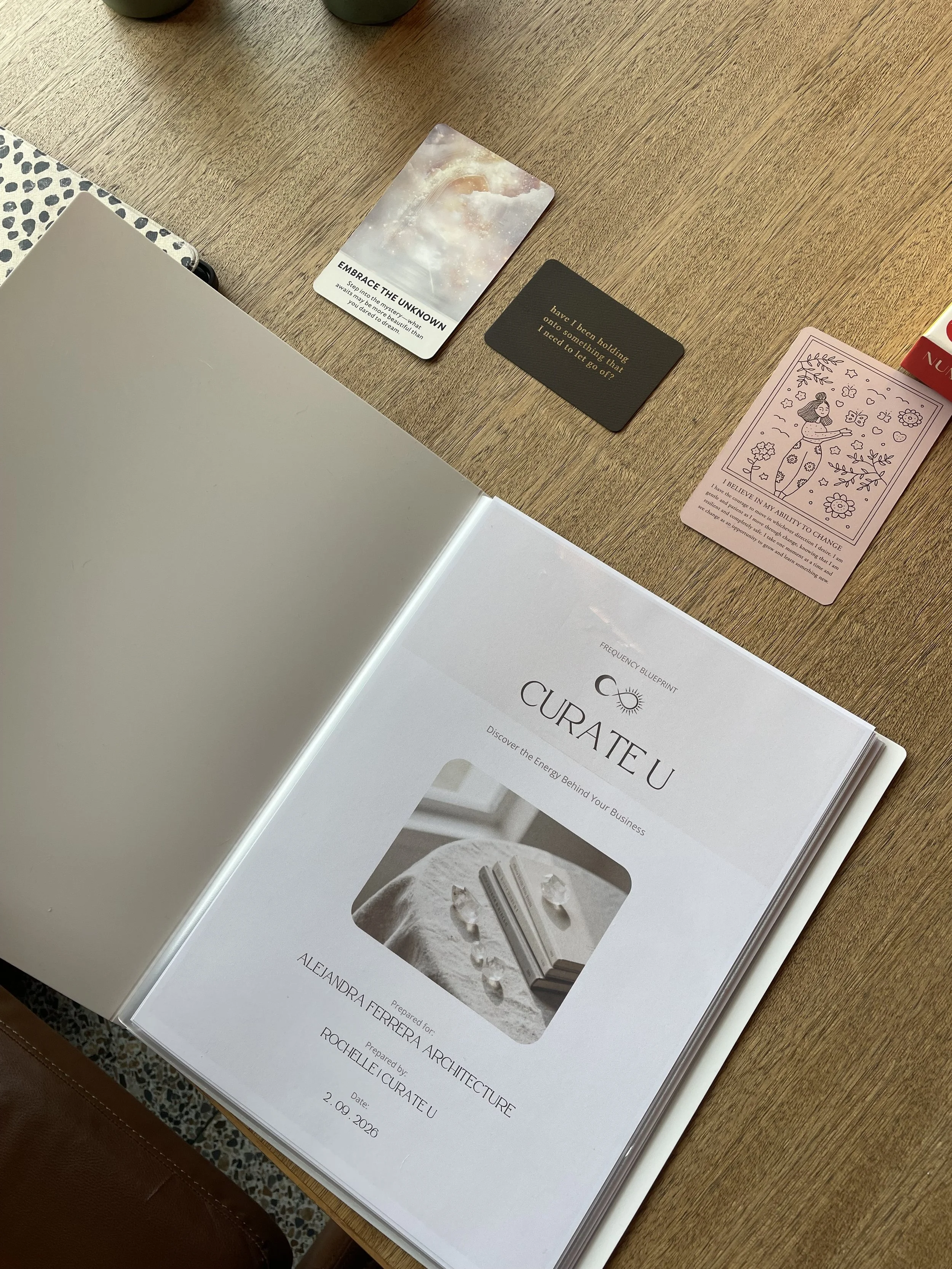 Open booklet with a cover page displaying the title 'CURA TE U' and a subtitle about discovering energy behind your business, along with a photo of crystals and a person. A few cards, including one with an abstract cosmic design labeled 'Embrace the Unknown' and another black card with a quote. A pink card with an illustration of a woman surrounded by flowers and a motivational quote about change. Part of a table and other objects are visible.