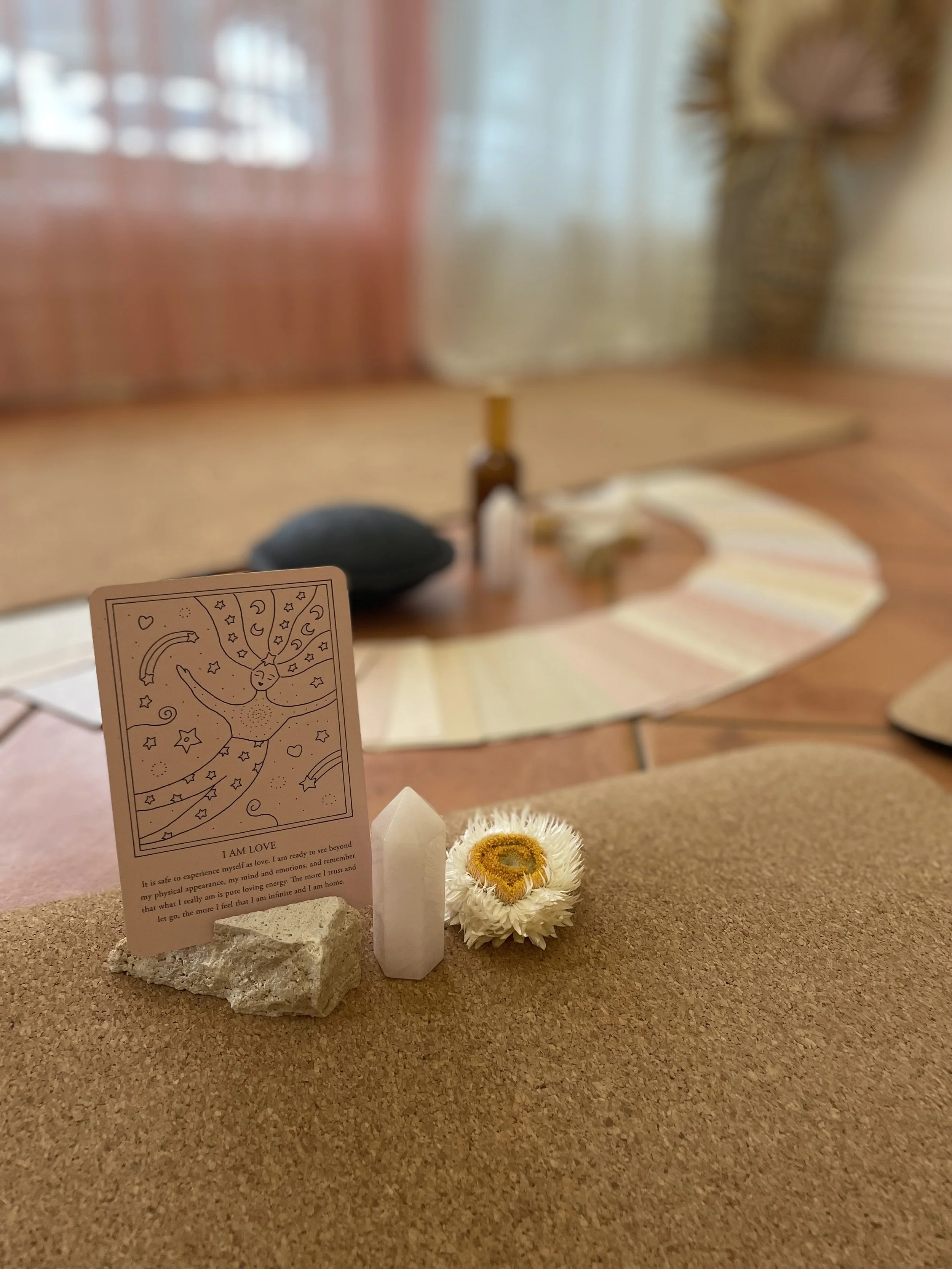 A close-up of a small altar on a kitchen table, featuring a card with a drawing of a person and the phrase "I AM LOVE," a white crystal, a dried white flower, and a small yellow object. In the background there is a blurred view of various other items and a plant.
