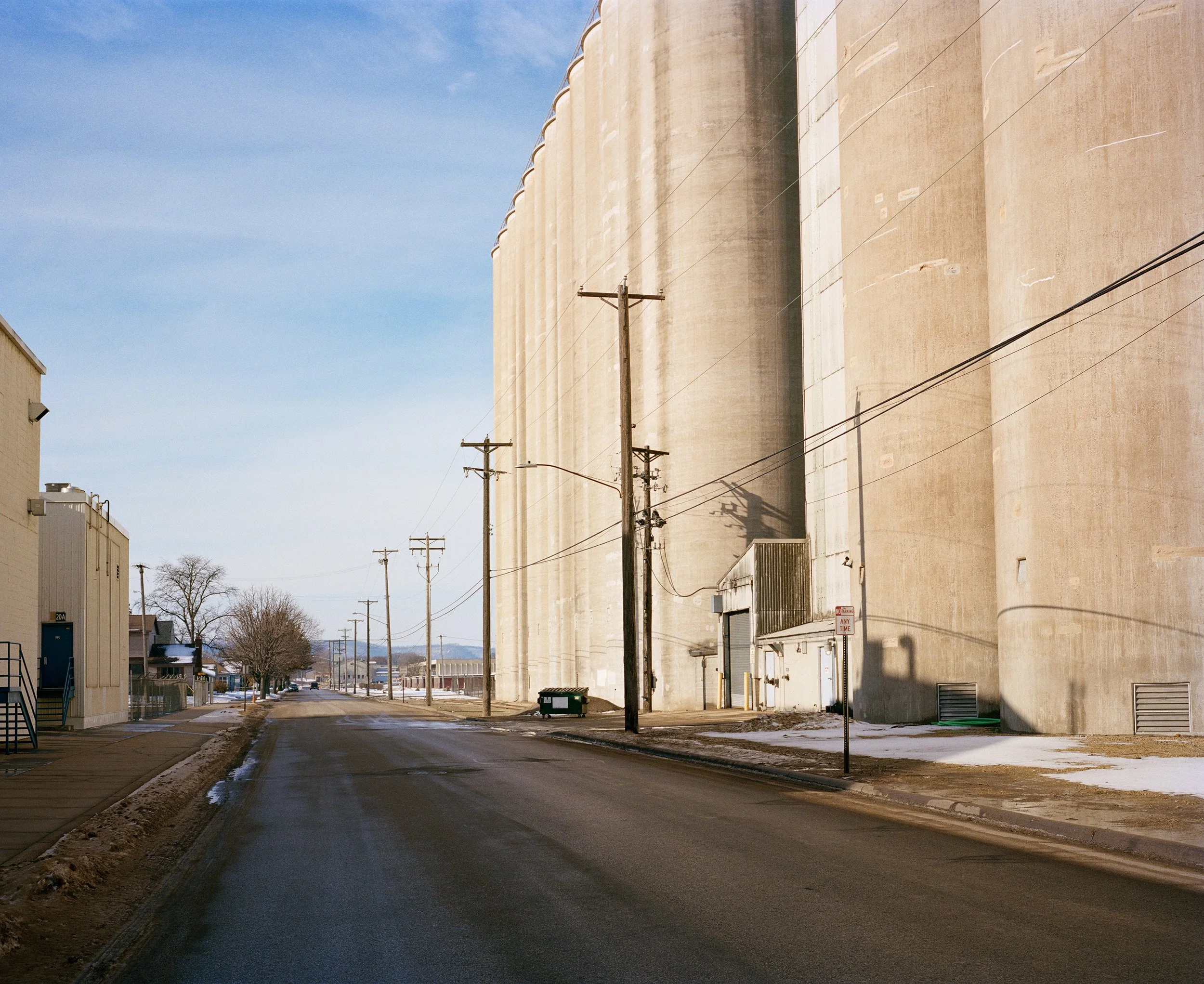 Empty street scene with large industrial grain silos on the right, utility poles along the sidewalk, snow on the ground, and a partly cloudy sky.