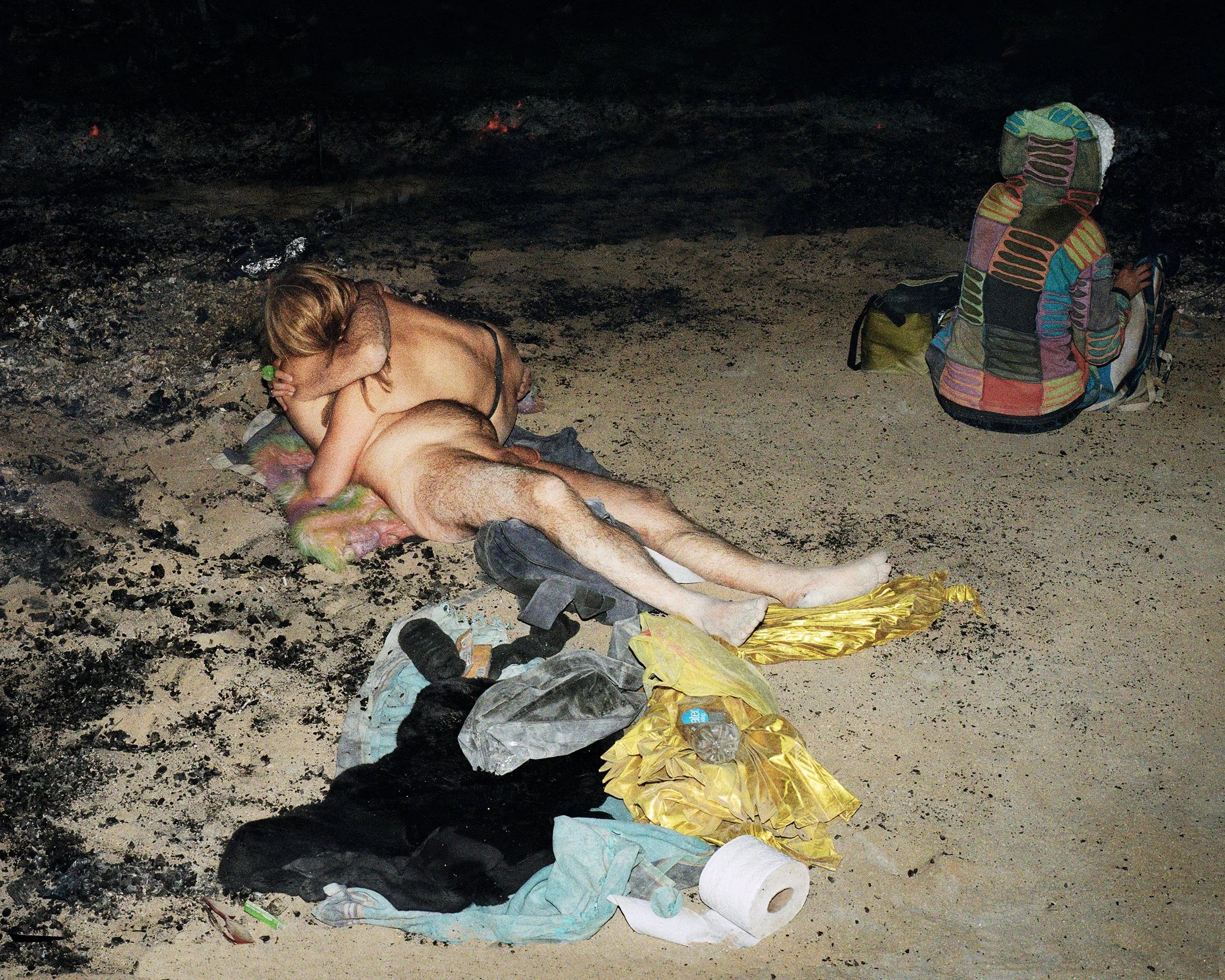 A man and woman lying on a beach at night, with discarded clothes and supplies around them, and a backpack nearby.