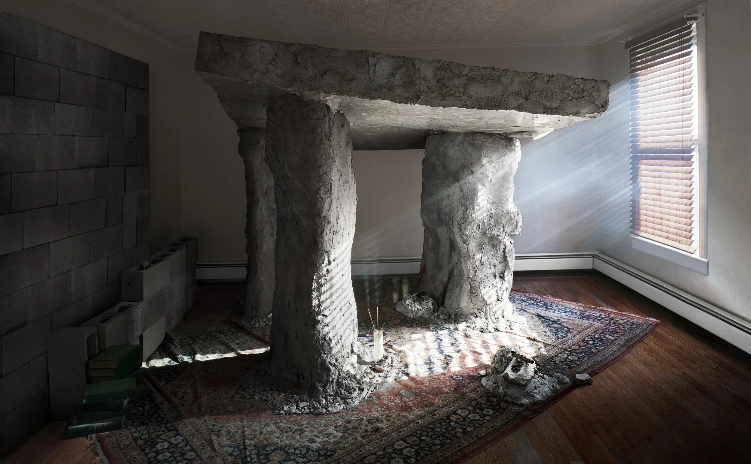 Interior of a room with three tall, irregularly shaped columns made of a stone-like material supporting a slab that serves as a tabletop. The room is lit by sunlight streaming in through a window with blinds, casting shadows on the floor and the colu