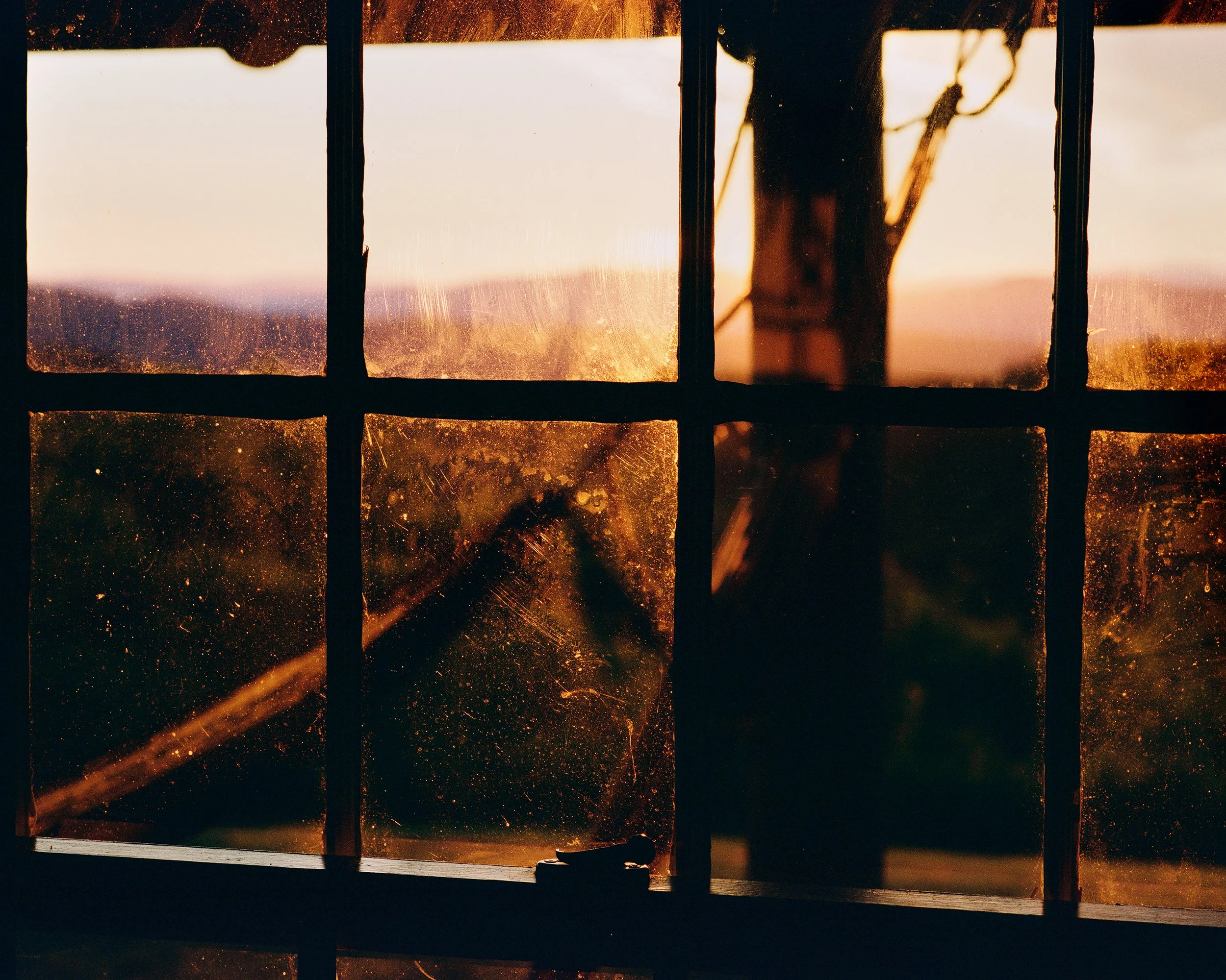 View of a sunset through a dusty, old window with grid panes, with a silhouette of a windmill in the background.