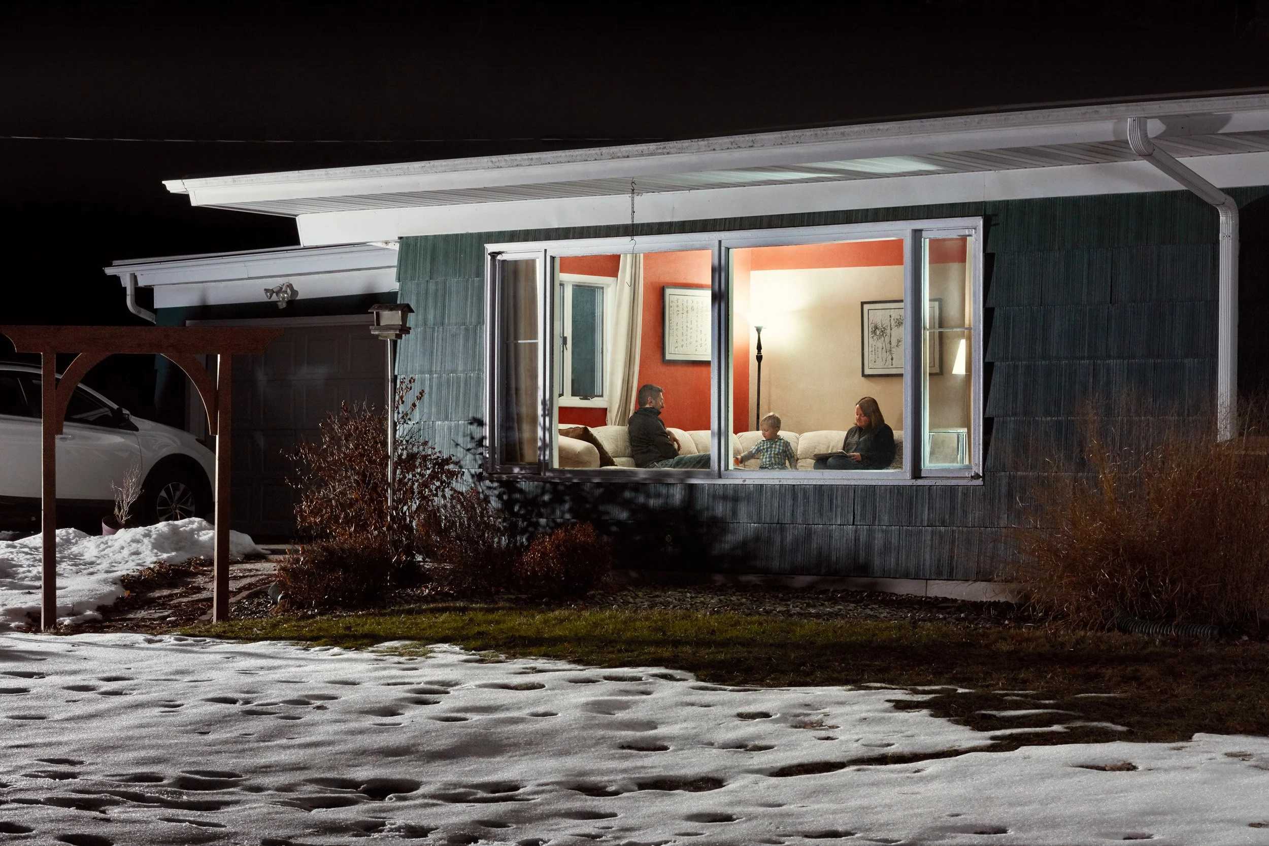 A house at night with illuminated interior viewed through large front windows showing three people sitting on a couch, including a man, a woman, and a child, inside a cozy living room.