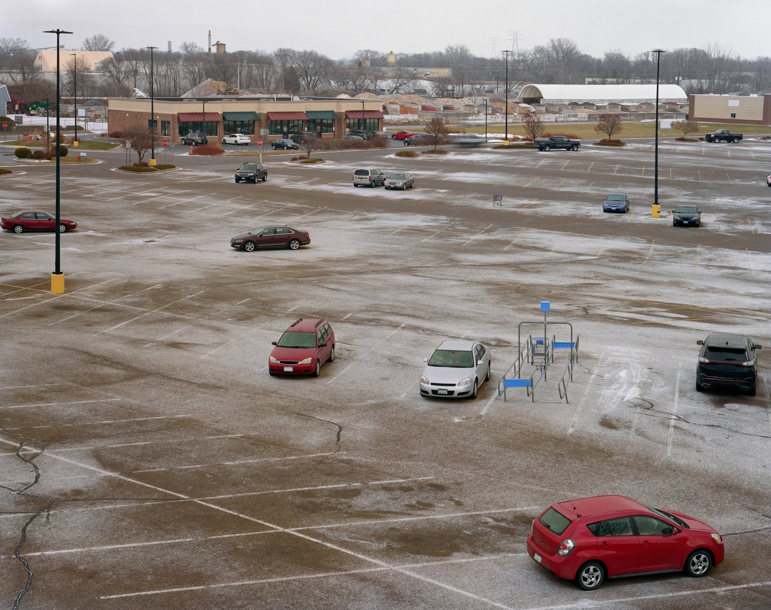 An empty parking lot with a few parked cars and shop buildings in the background, on a cloudy winter day.