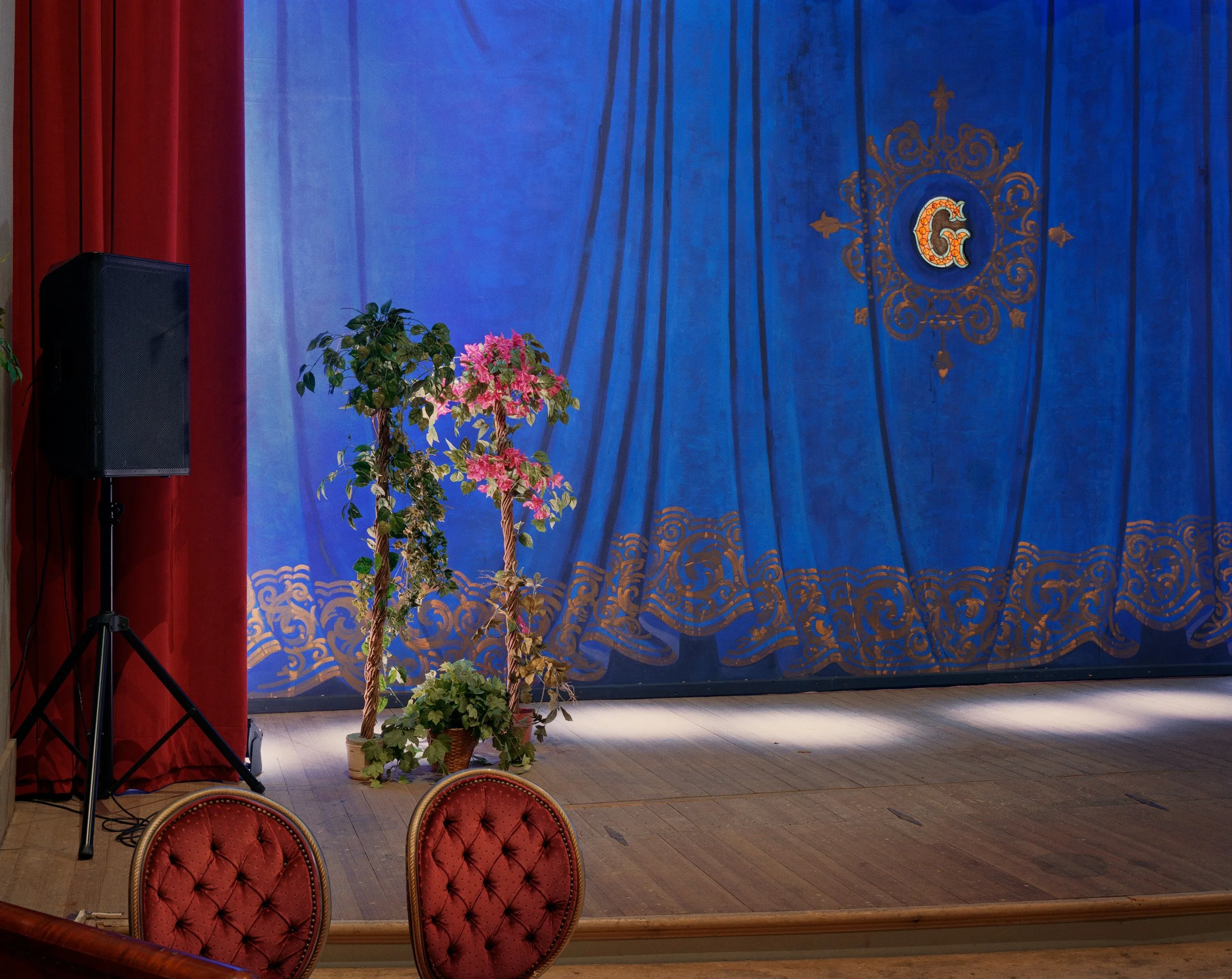 A stage with closed blue curtains decorated with a gold letter G and ornate patterns, red velvet chairs in the foreground, two potted plants on the left side, and a black speaker on a stand.