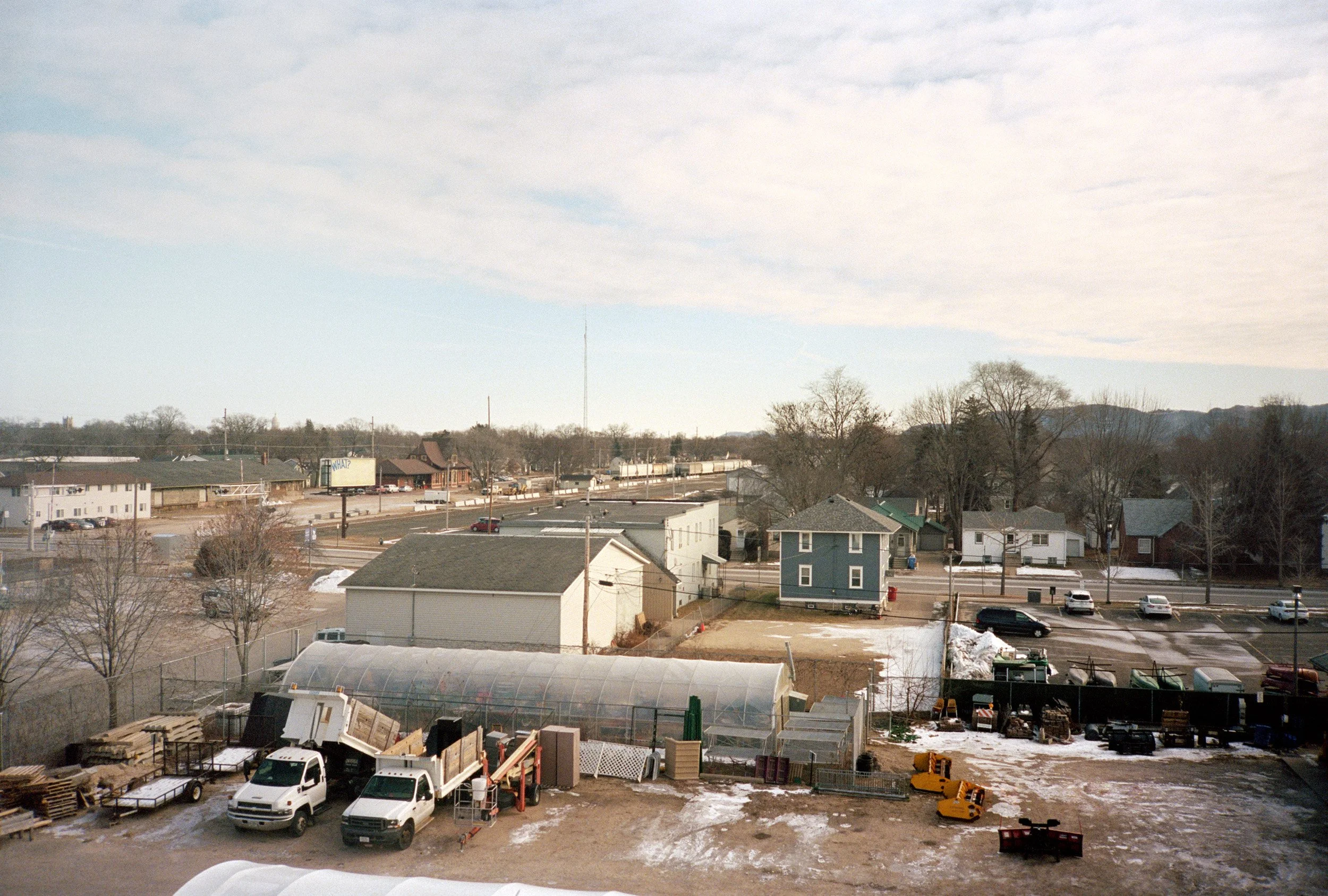A suburban neighborhood with houses, trees, and a parking lot with multiple cars, winter scenery with some snow, and a clear sky.