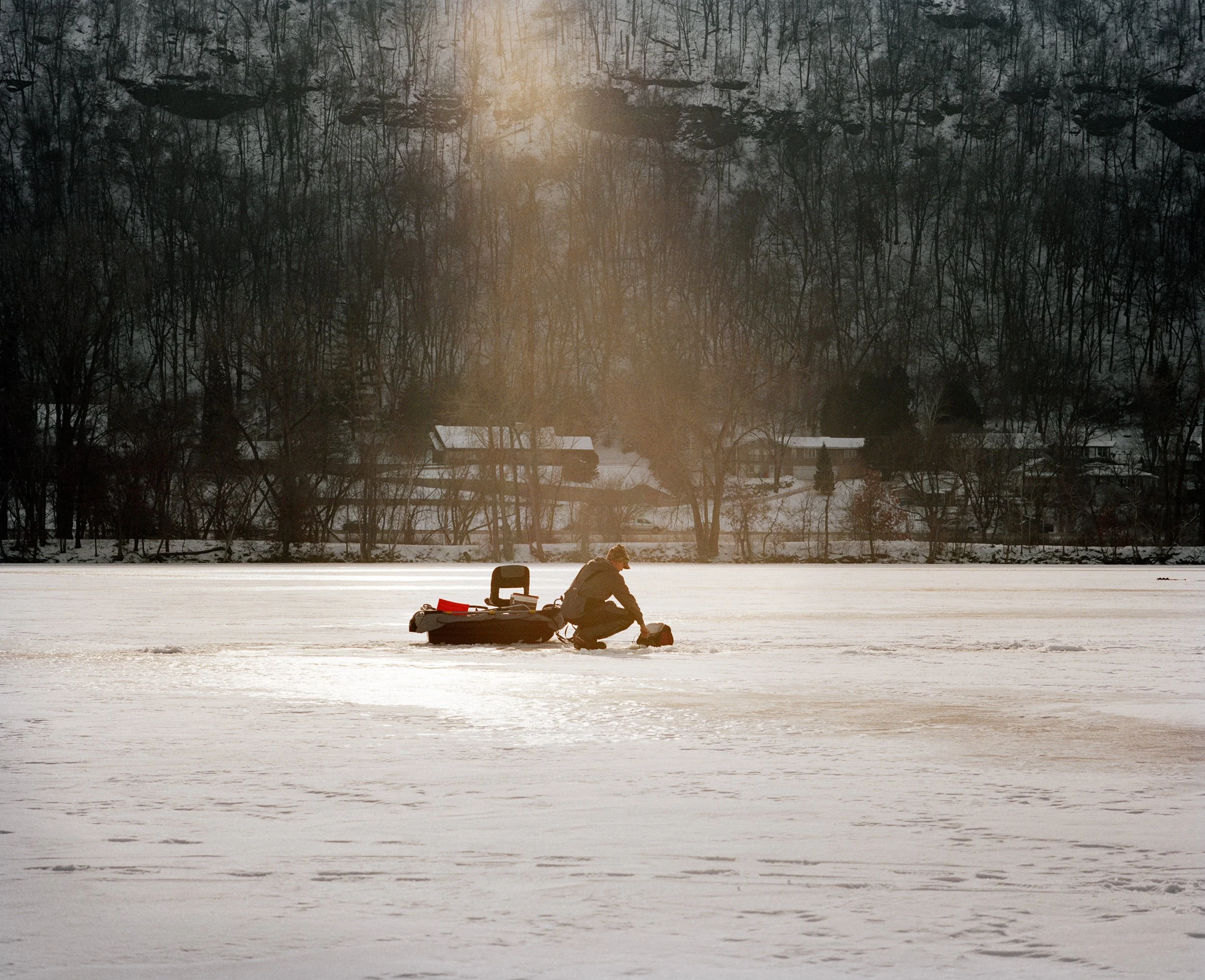 A person ice fishing on a frozen lake during sunset, with a cabin and trees in the background.