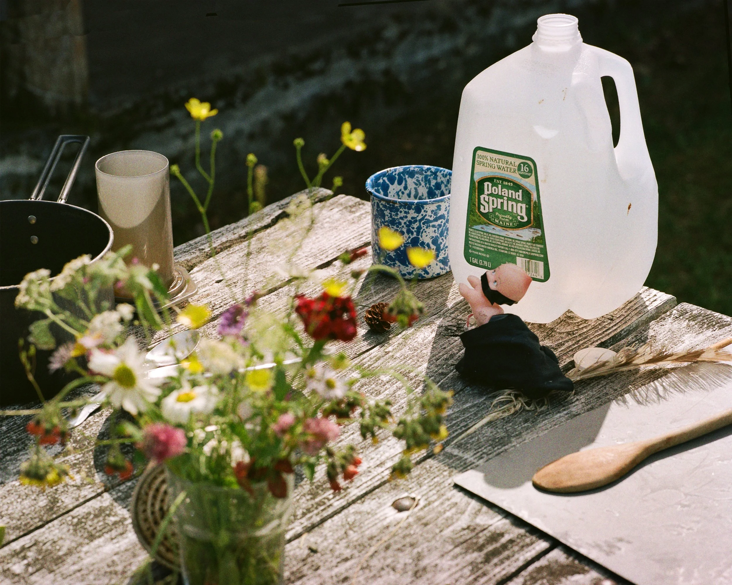 A rustic wooden outdoor table holding a vase of mixed wildflowers, a black pan, a beige cup, a blue speckled mug, a large plastic jug of water, a small pine cone, a doll with a black dress, a feather, and a wooden spoon, with a grassy background.