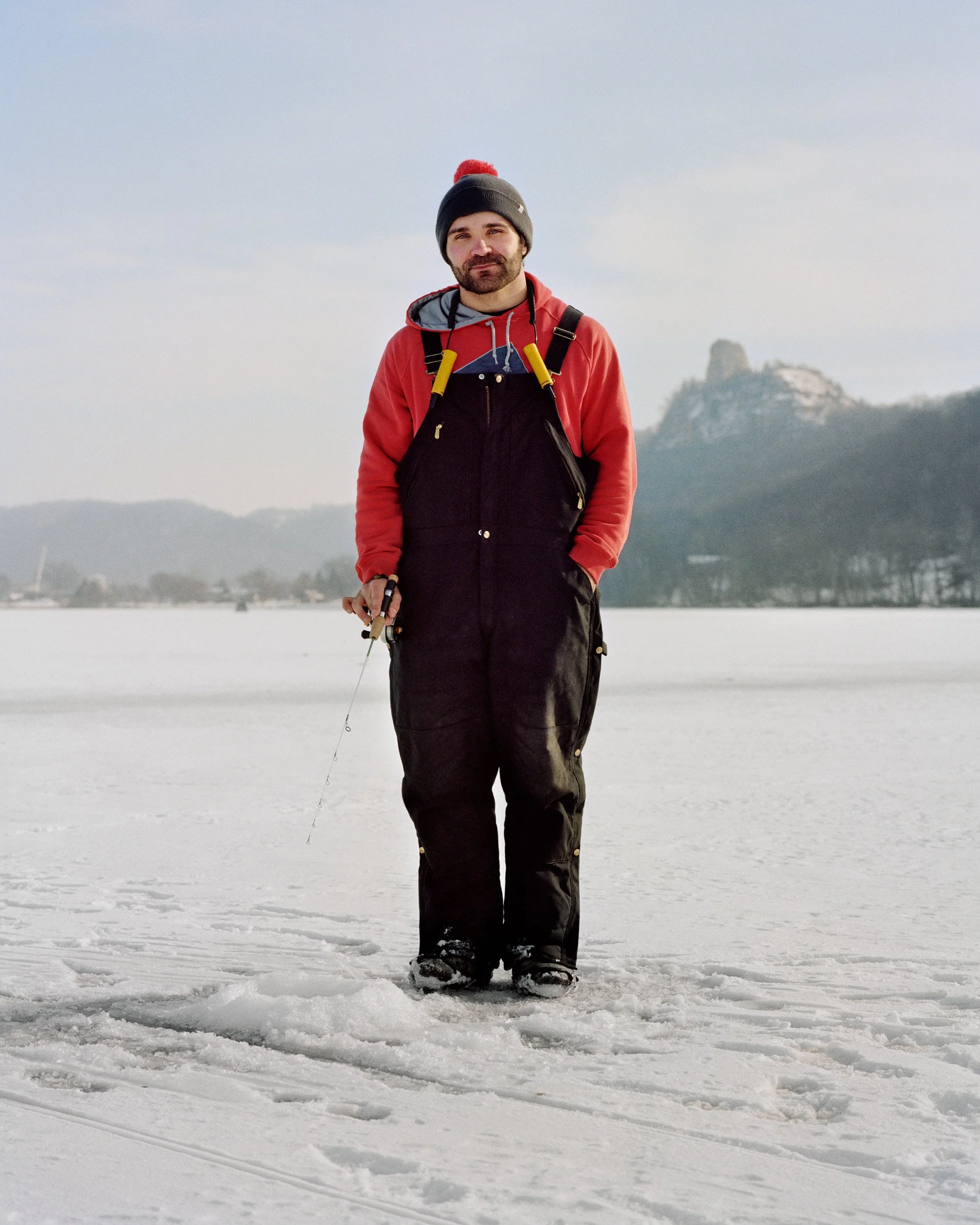 Man standing on snow-covered lake holding a fishing rod, dressed in winter clothing, with a mountain in the background.