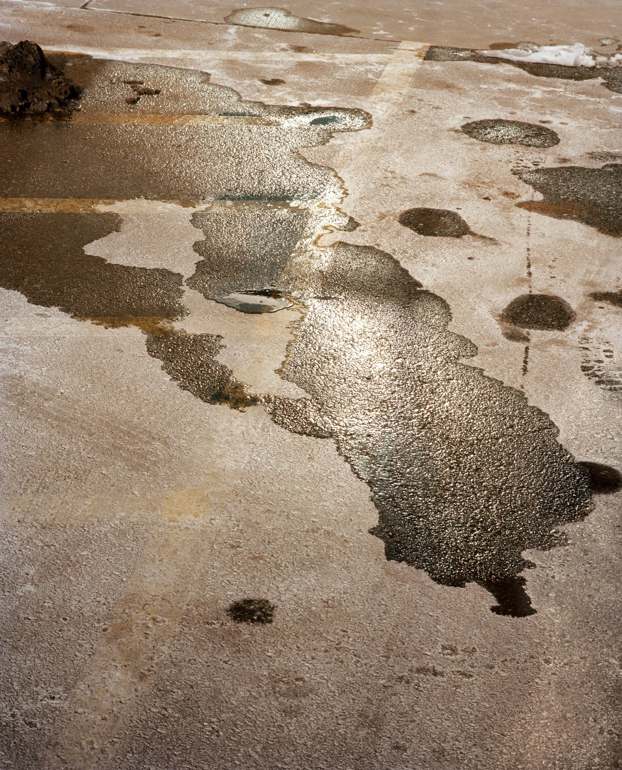 Wet patches and ice on pavement reflecting sunlight, with some small puddles and water streaks.