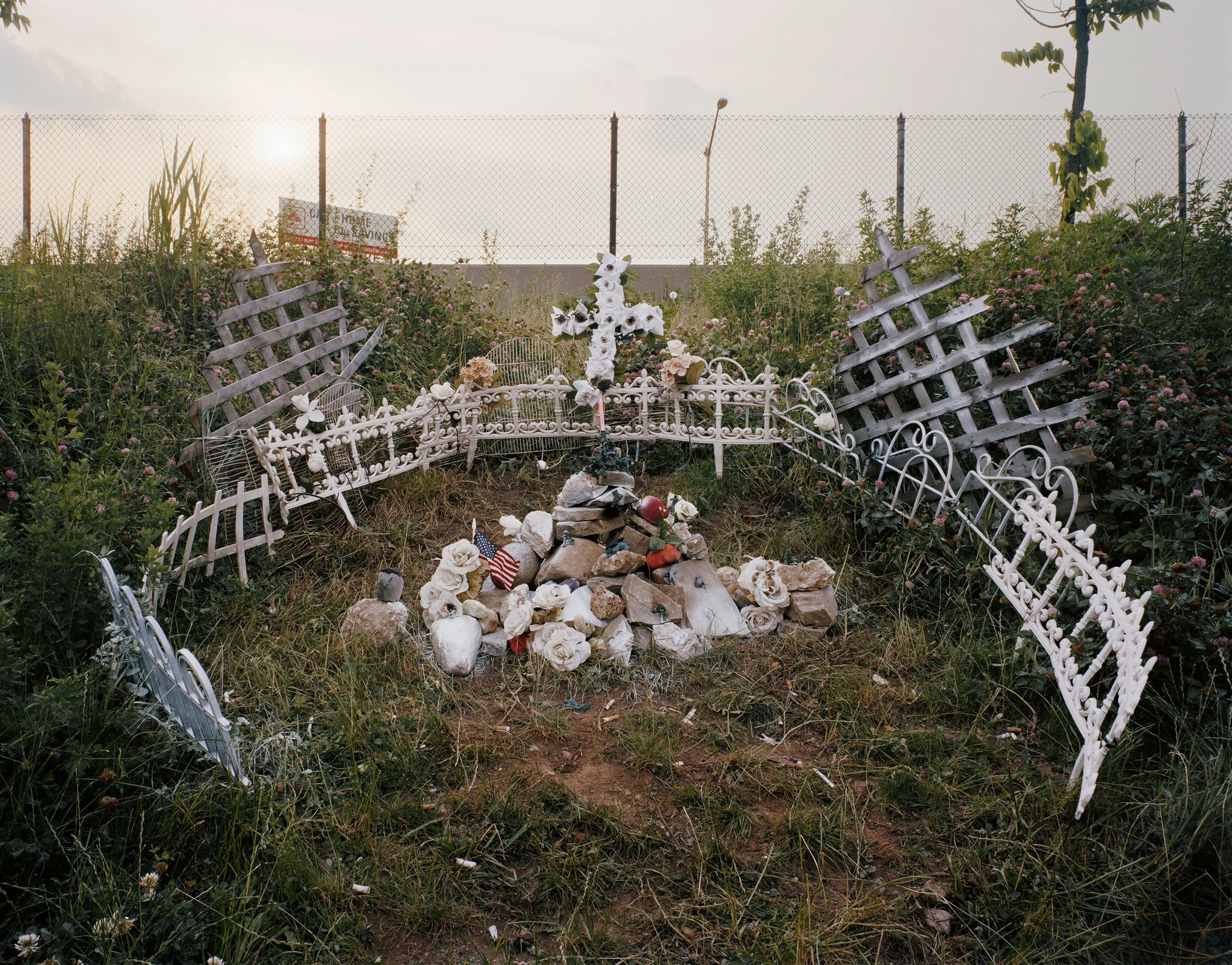 A small outdoor memorial with a white cross, flowers, and rocks, surrounded by broken white metal fences and overgrown grass, with a chain-link fence and a billboard in the background.