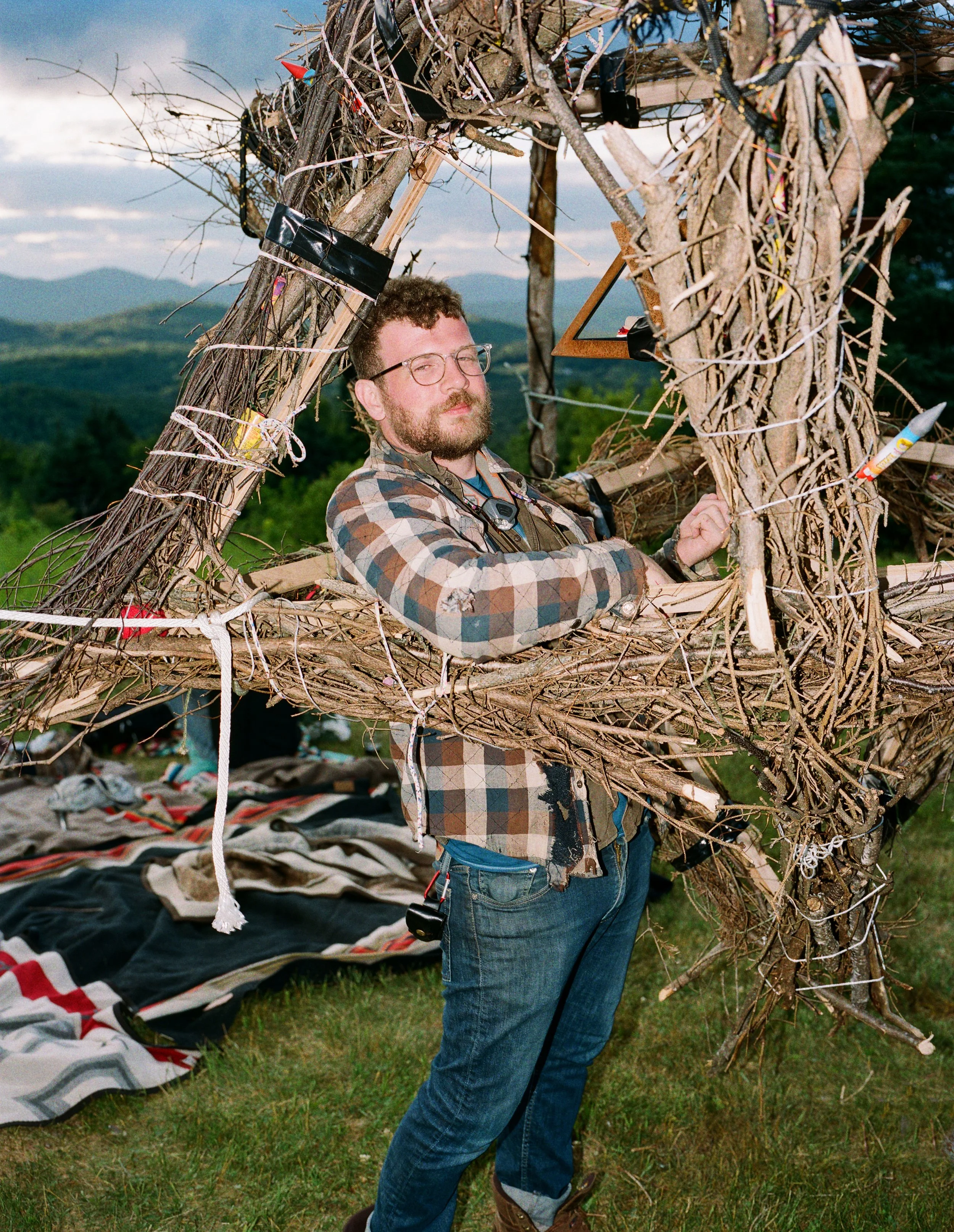 A man with glasses and a beard wearing a plaid shirt and jeans, carrying a large handmade wooden and twig structure outdoors in a grassy field, mountains in the background, with blankets on the ground.