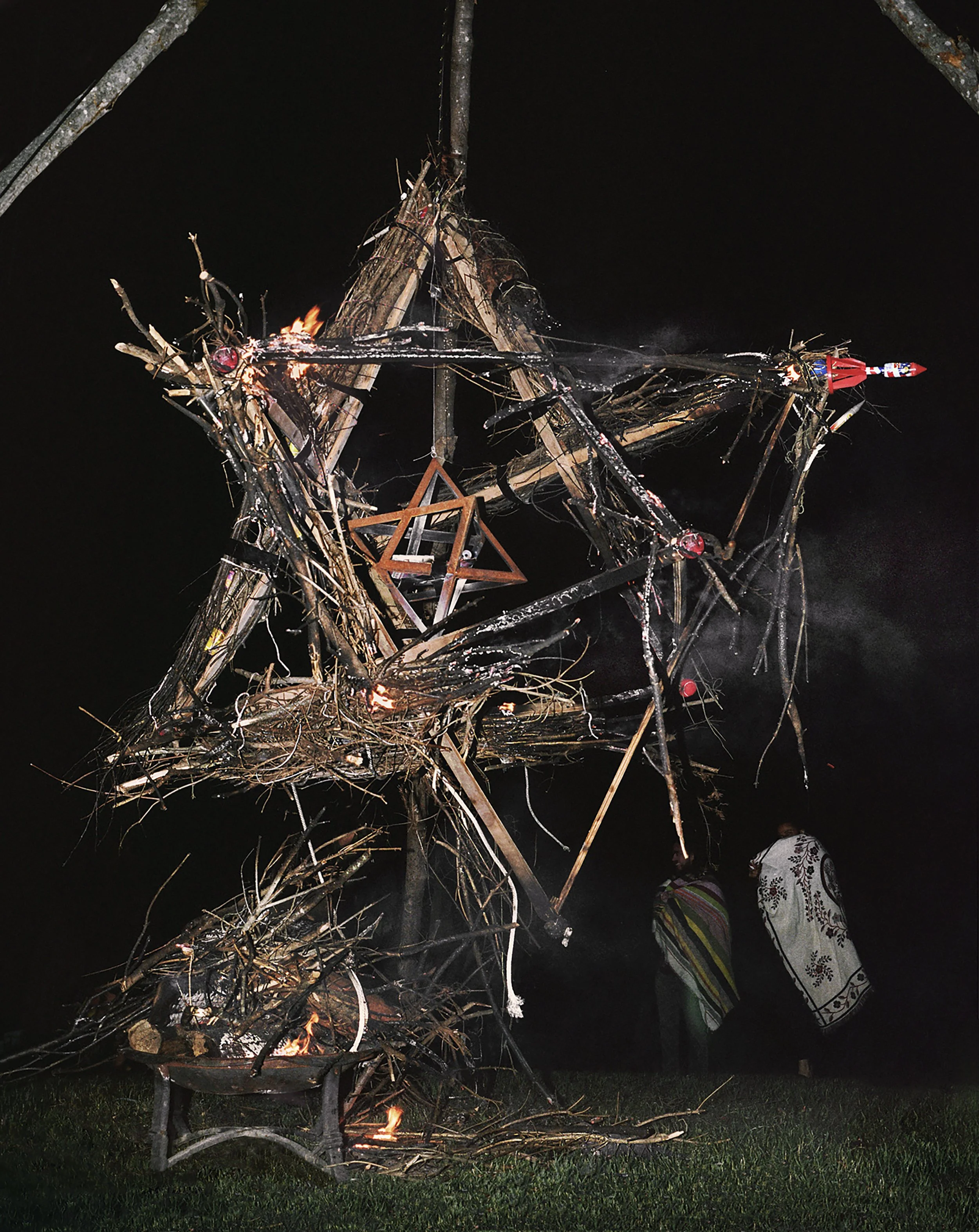 A large bonfire made of wooden sticks and logs with flames burning at the base. The fire forms a star shape with a wooden Star of David hanging in the middle. Two people are standing nearby with blankets, one striped and the other with a white floral