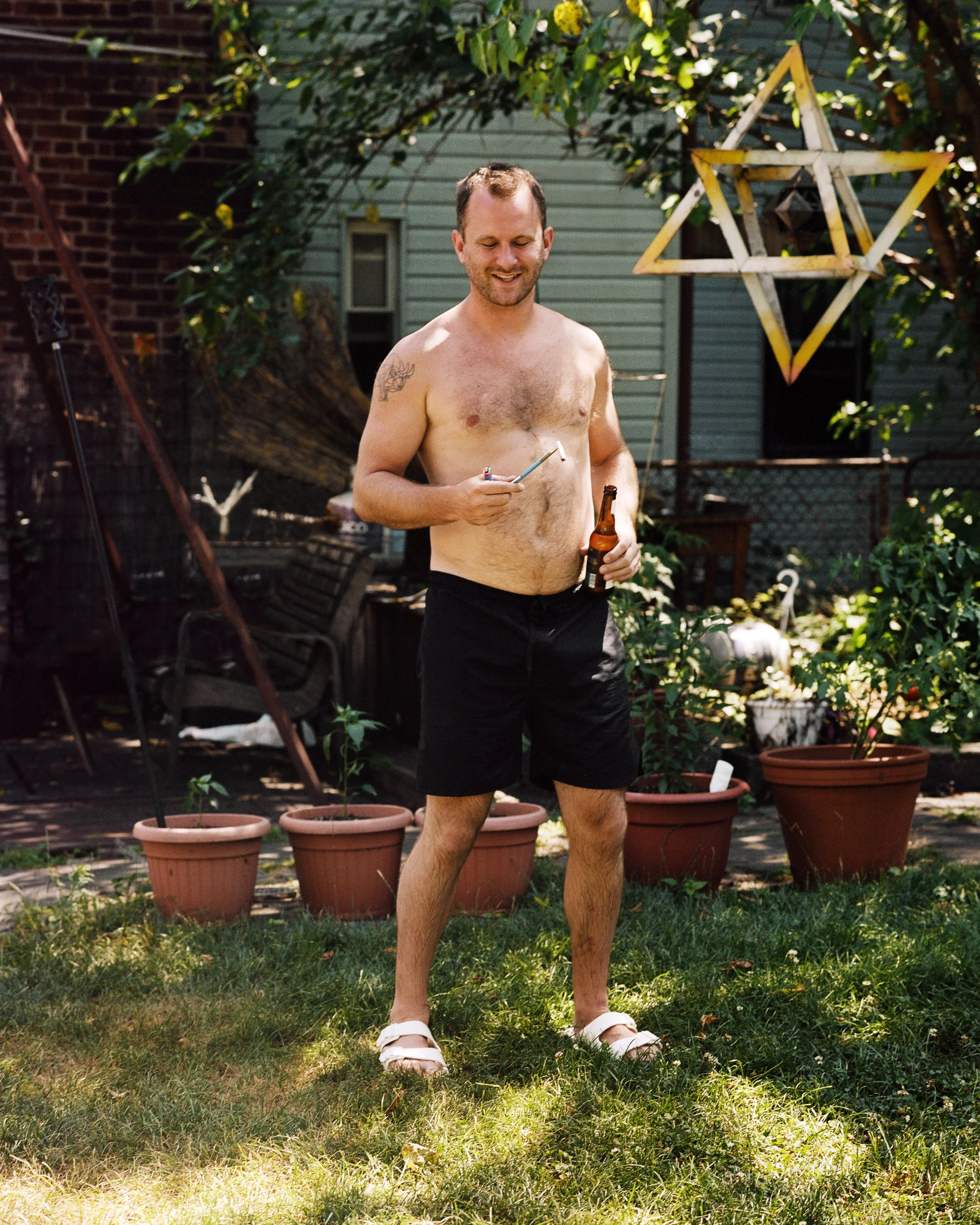 A shirtless man holding a beer bottle and a lit cigarette standing on grass in a backyard with potted plants and a wooden star decoration hanging from tree branches.