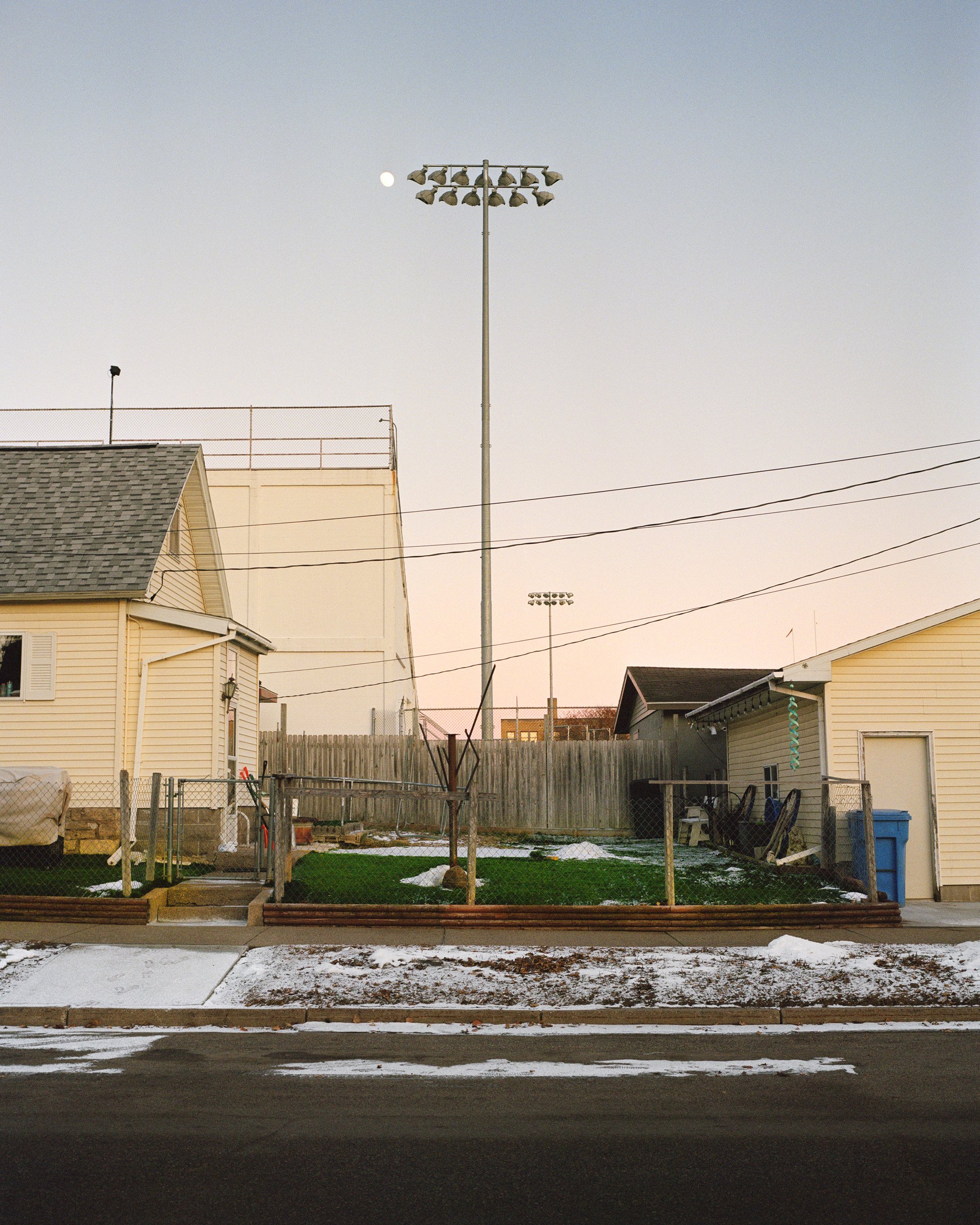 Residential backyard with a fence, grass, two small houses, and large baseball field lights in the background with a visible partial moon in the sky.