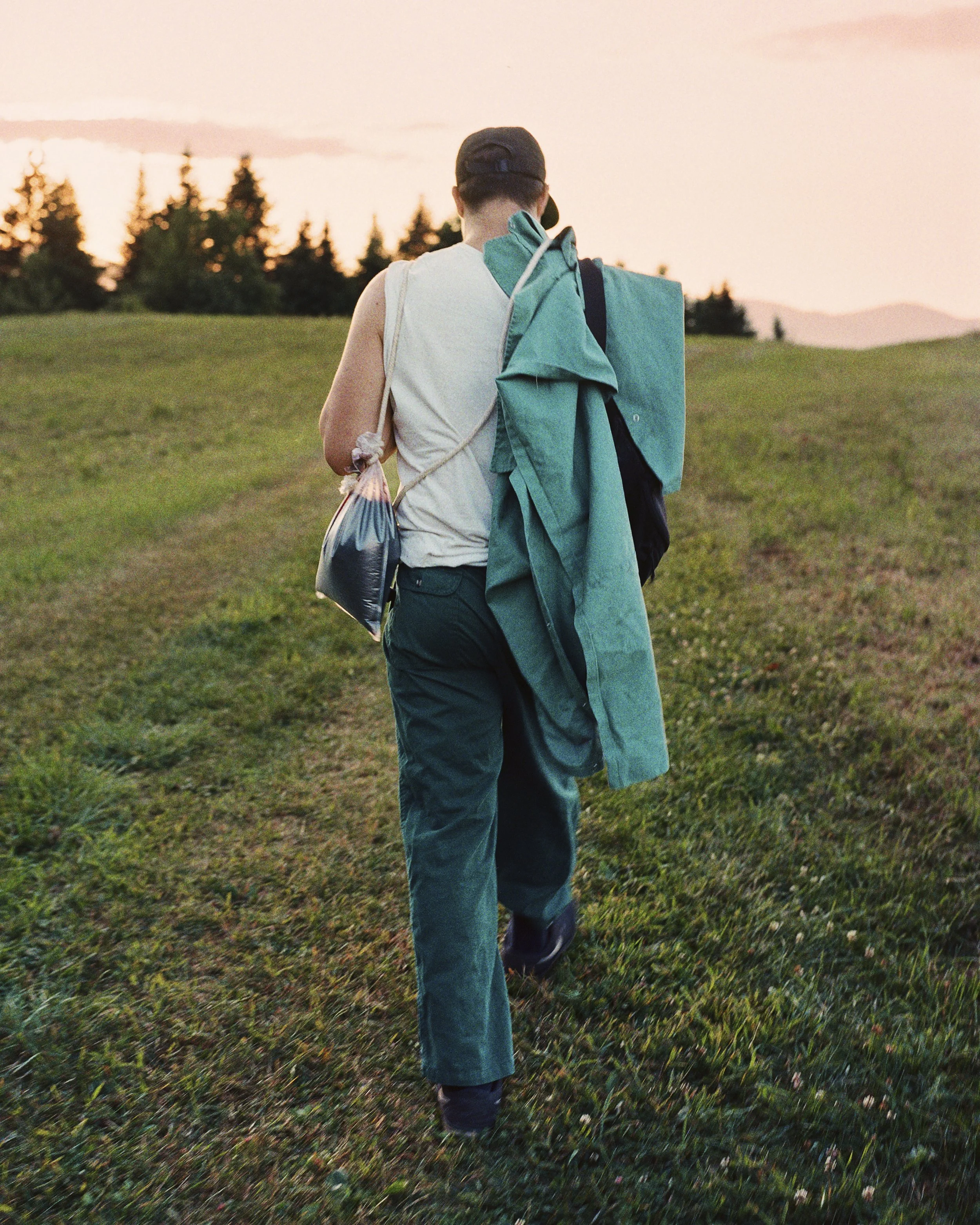 A person walking through a grassy field at sunset, carrying camping gear including a large blue-green backpack and a blanket, with trees and mountains in the background.
