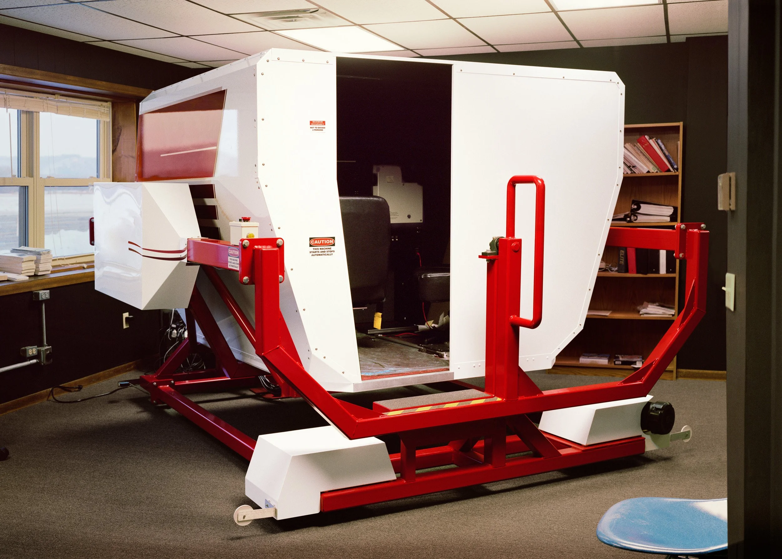 A large white and red machine resembling a flight simulator sits in a room with bookshelf and window.