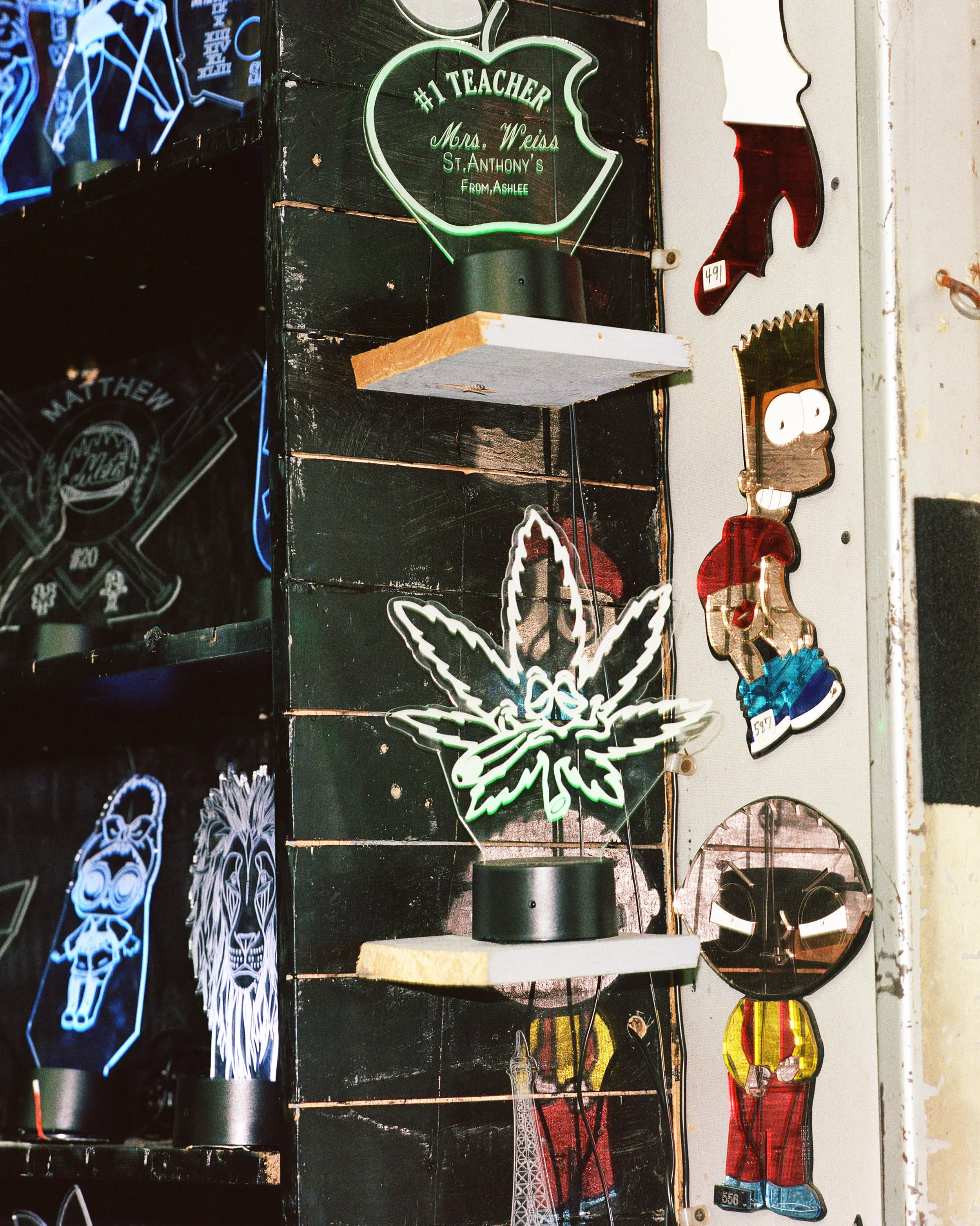 Collection of colorful neon and acrylic signs with various designs, including a green leaf with text, cartoon characters, and abstract patterns displayed on a wall.
