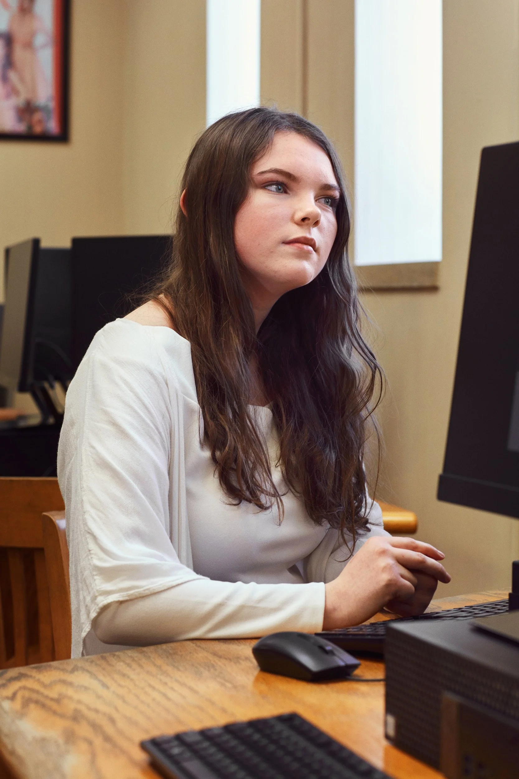 A young woman with long, wavy brown hair working on a desktop computer in an office setting.