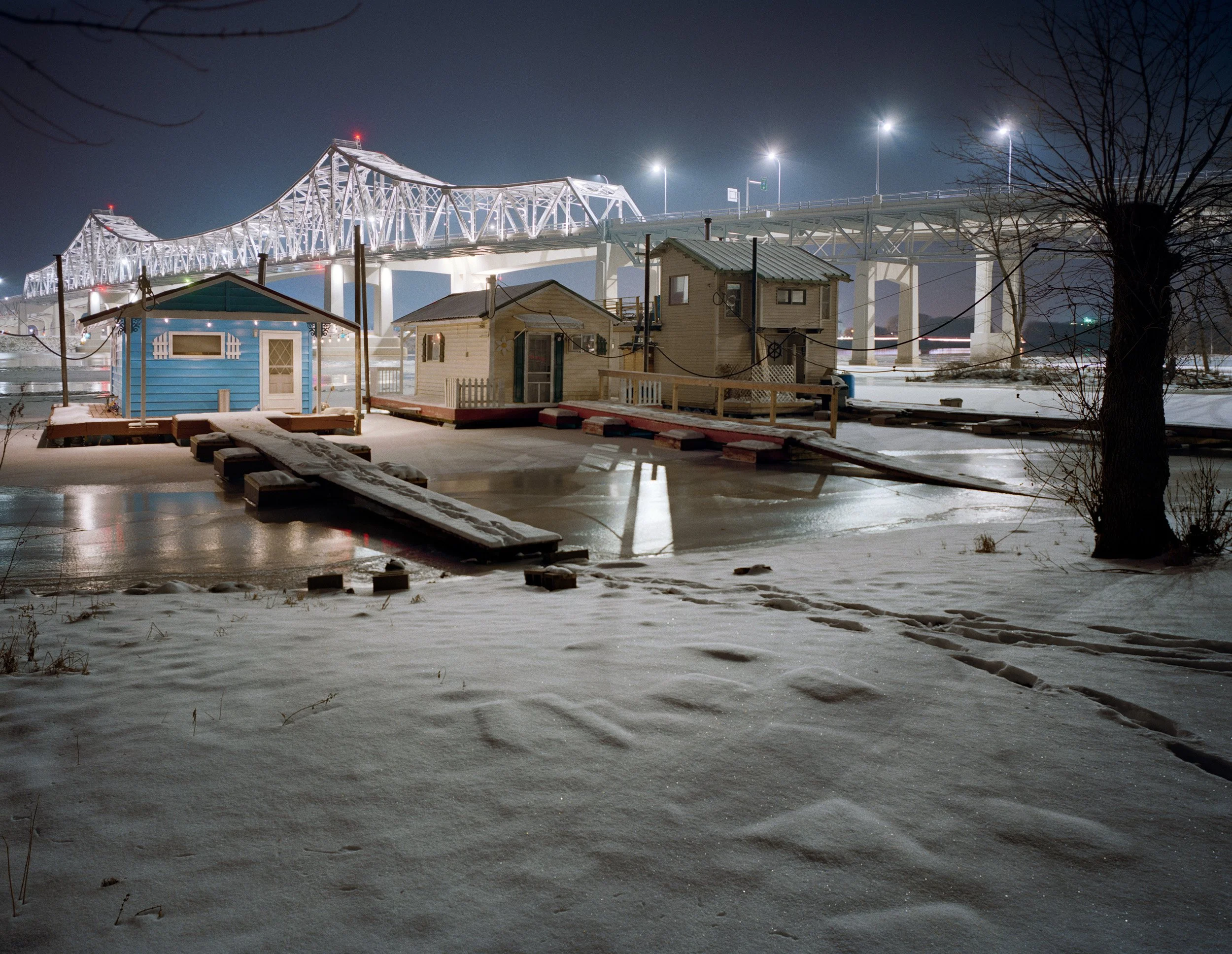 Nighttime scene of small colorful houses on a snowy, icy waterfront with a large illuminated bridge in the background.