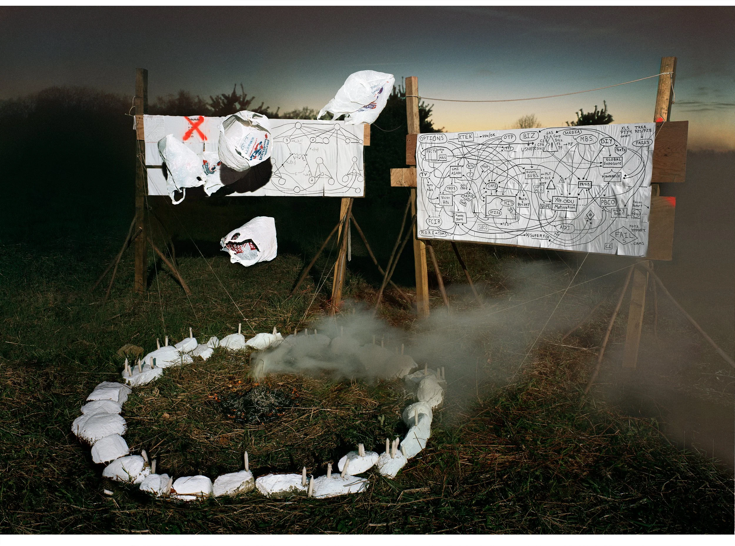An outdoor scene featuring two large whiteboards with complex diagrams and notes, several crumpled paper bags hanging on the left, a circle of rocks with small candles in the foreground, and a foggy atmosphere during sunset.