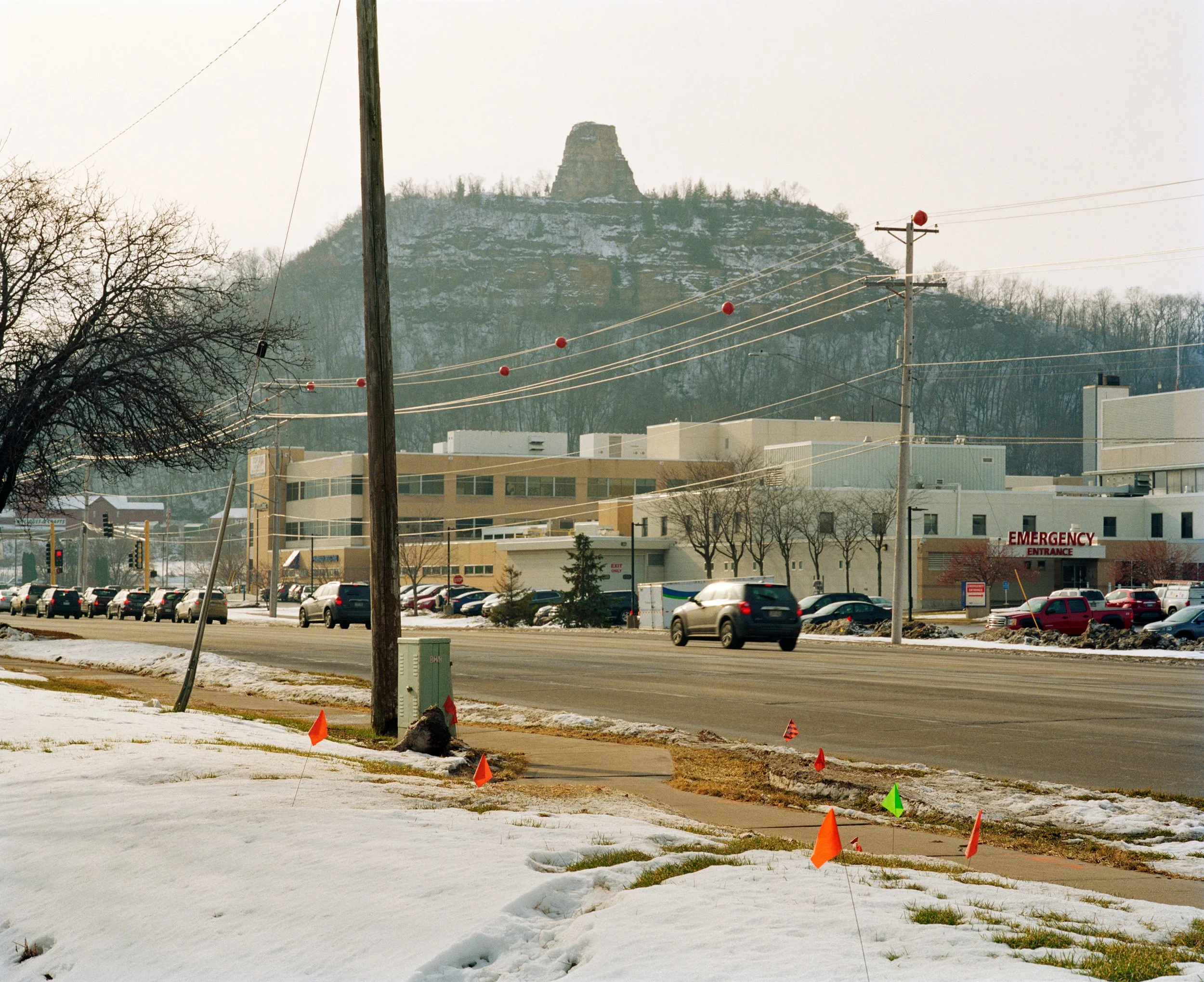 Street view with emergency entrance of a hospital, parked cars, power lines, and a mountain in the background during winter.