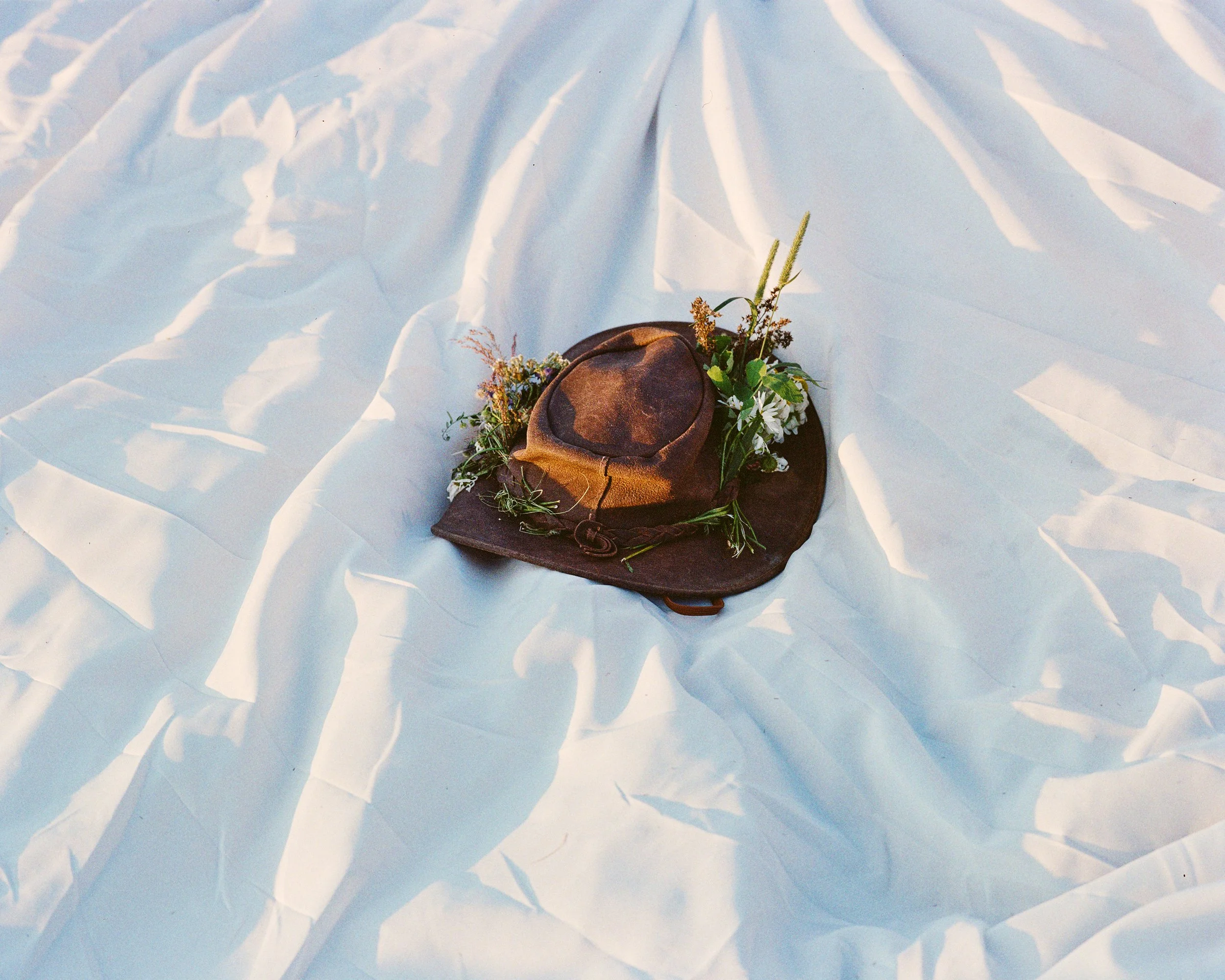 A brown cowboy hat decorated with flowers and greenery, placed on a white, ruffled fabric background.