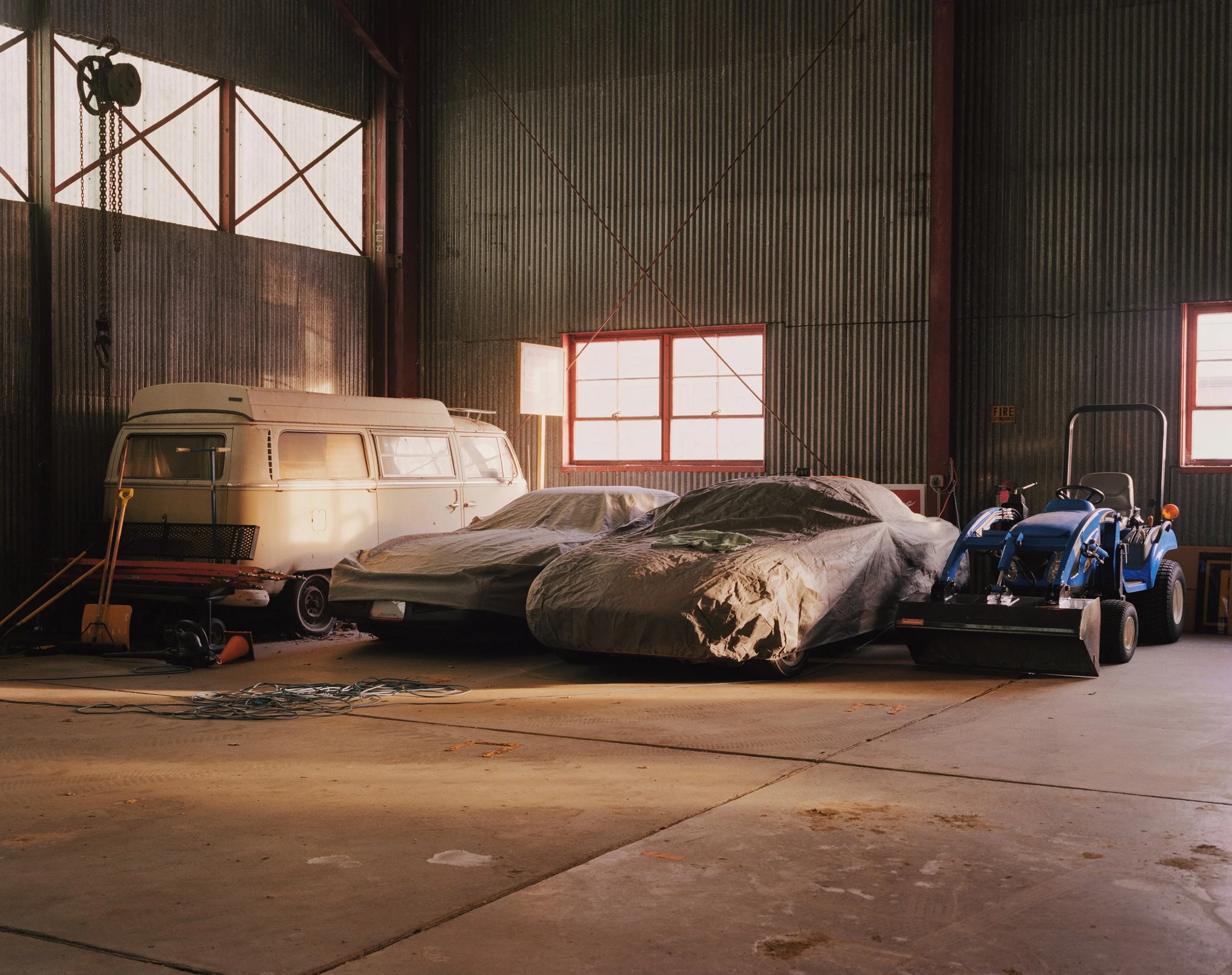 Inside a garage with two covered cars, a vintage van, and a blue riding lawn mower, with industrial metal walls and red window frames.