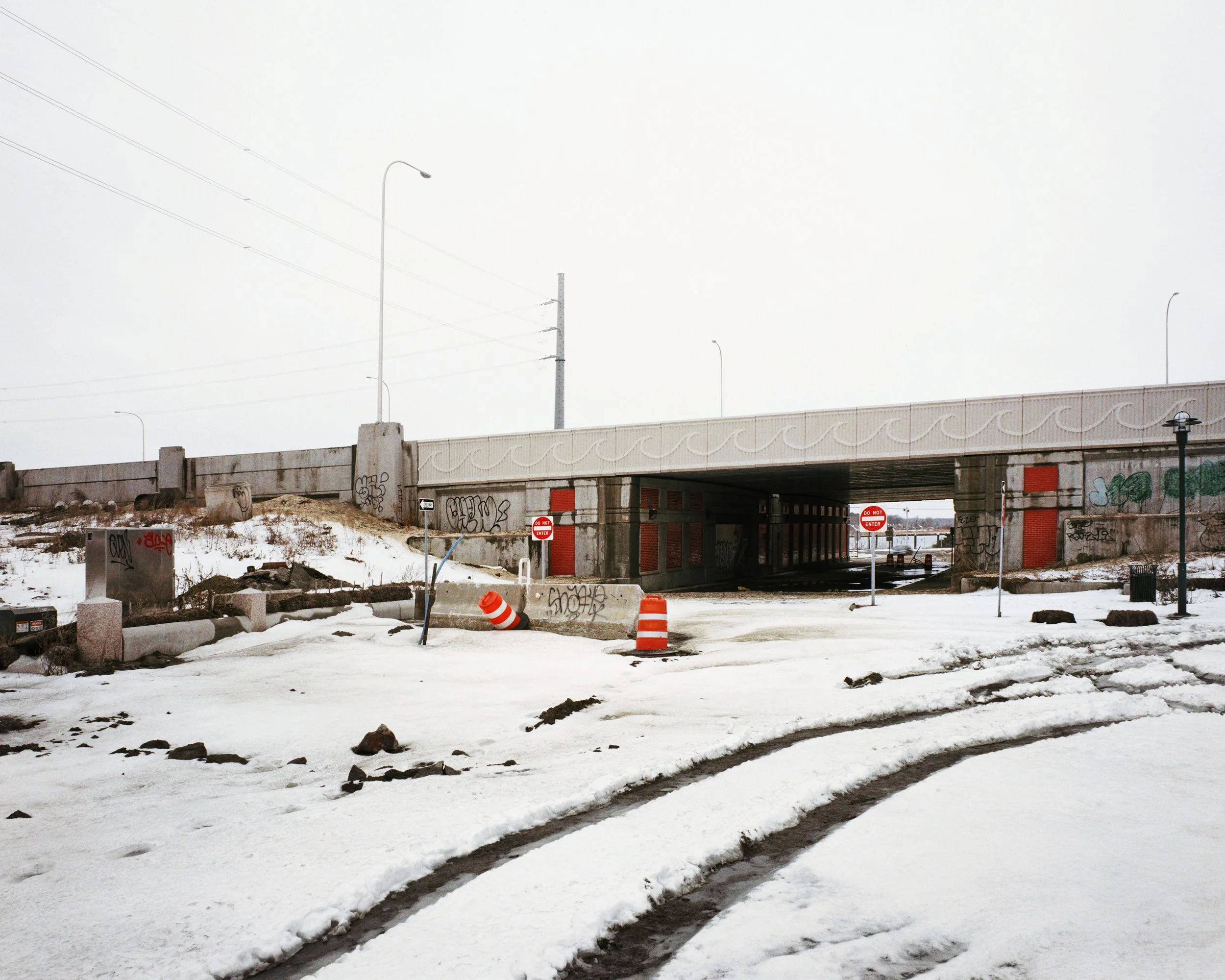 Snow-covered area with an underpass or bridge, graffiti on the walls, traffic signs including 'Do Not Enter', and orange traffic cones, with a foggy or overcast sky.