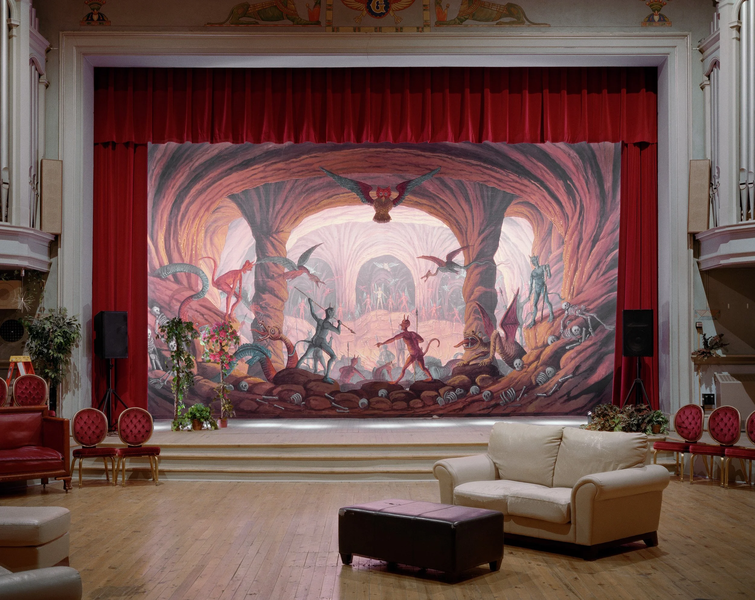 Theater stage with mural depicting a fantasy scene of dragons, demons, and monsters in a cave with red curtains on sides and a few chairs and couches in front.