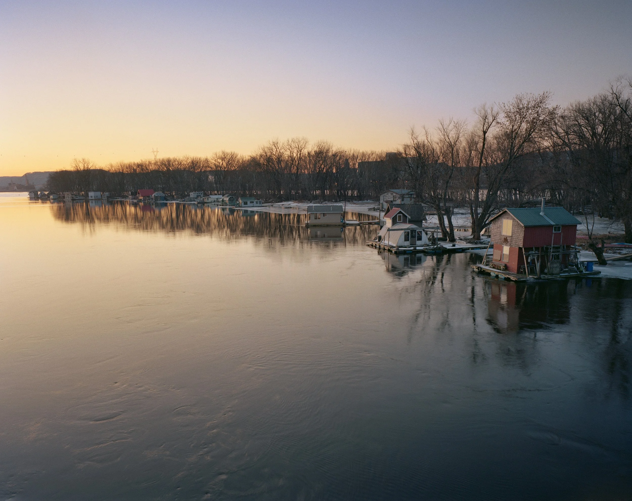 Dusk over a calm river with floating houses, some with decks, and leafless trees along the shoreline.