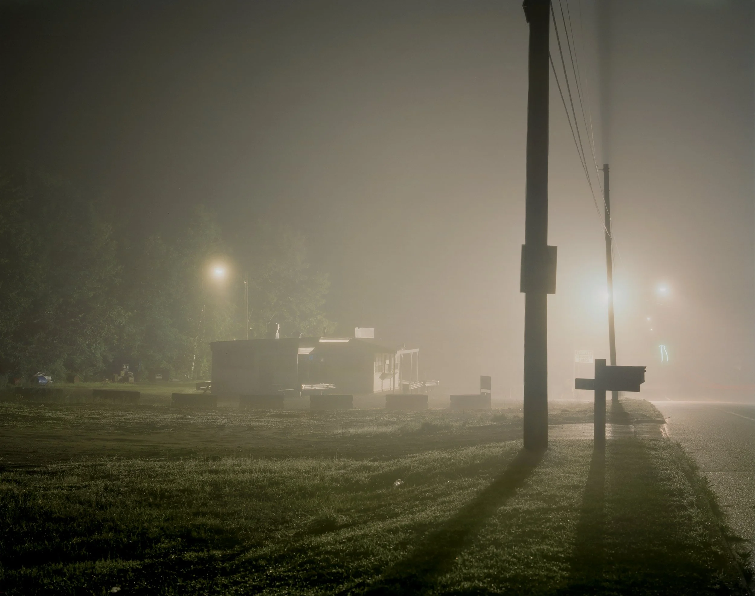 A foggy night scene with streetlights illuminating a grassy area, utility poles casting long shadows, and a small building or trailer in the background.