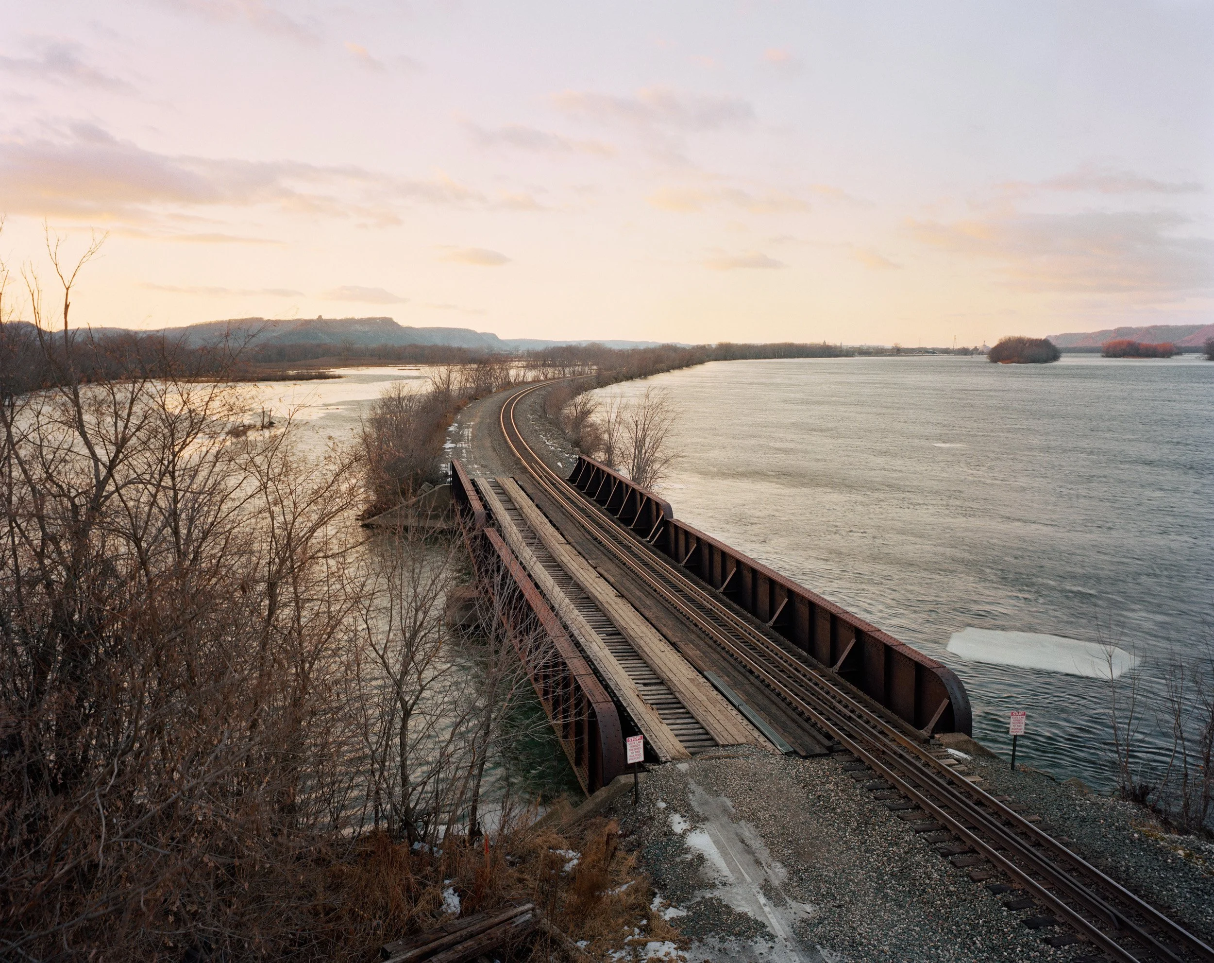 A railroad bridge crossing over a body of water with trees on either side, and a distant landscape with hills under a pastel sky.