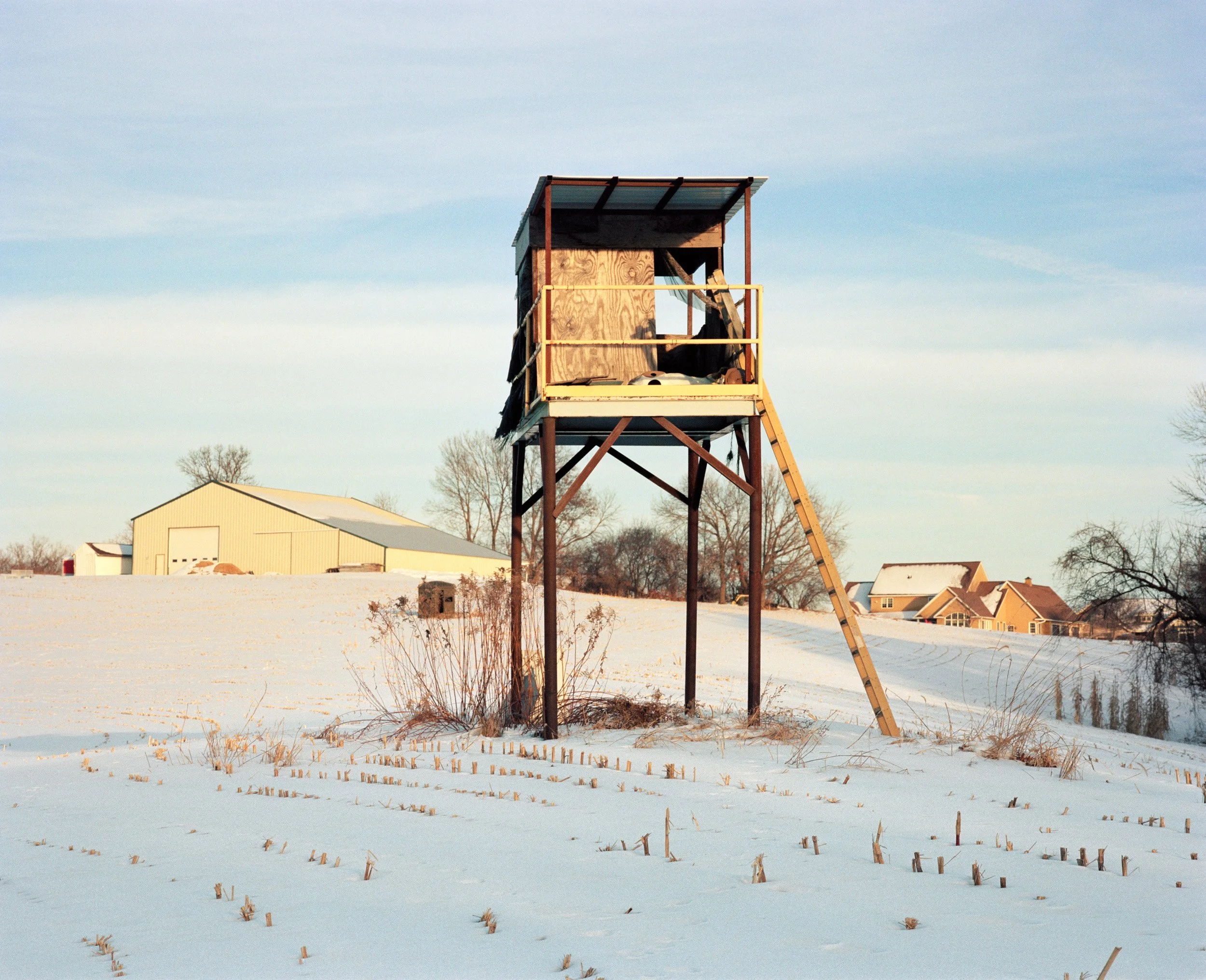A wooden hunting or observation tower in a snow-covered field with a clear sky, a few trees, and houses in the background.