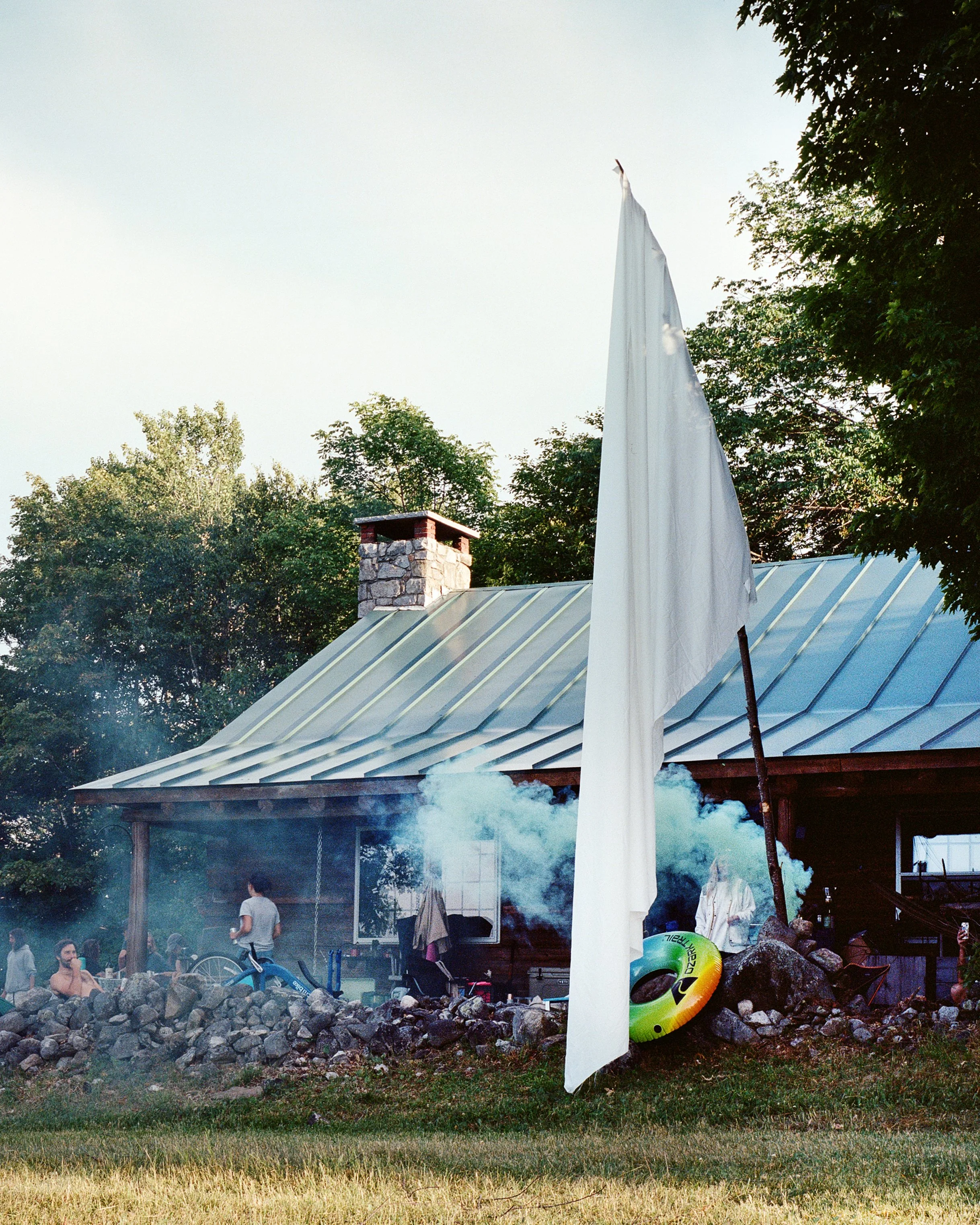 People gather outside a house with smoke rising from it, a large white flag in the foreground, a floatation ring on rocks, and trees in the background.