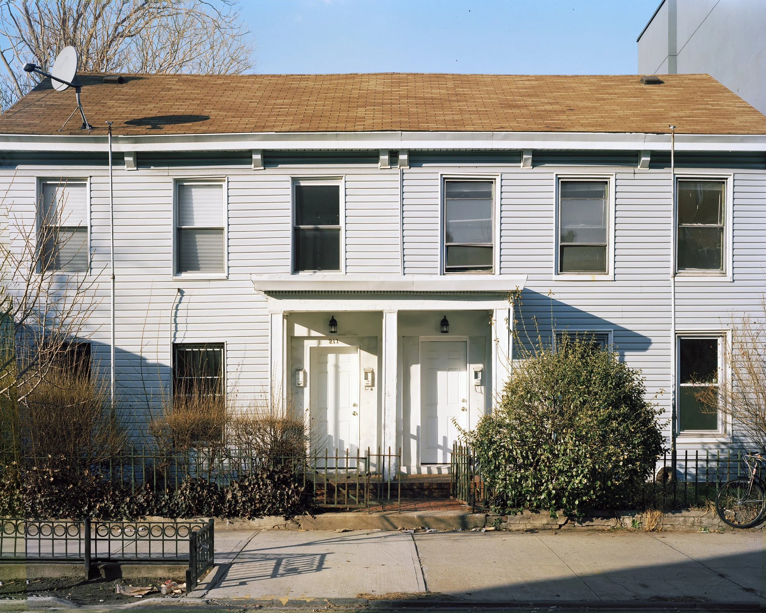 A two-story residential building with white siding, multiple windows, and two front doors. There are bushes and a small fence in front, with a bicycle leaned on the right side.