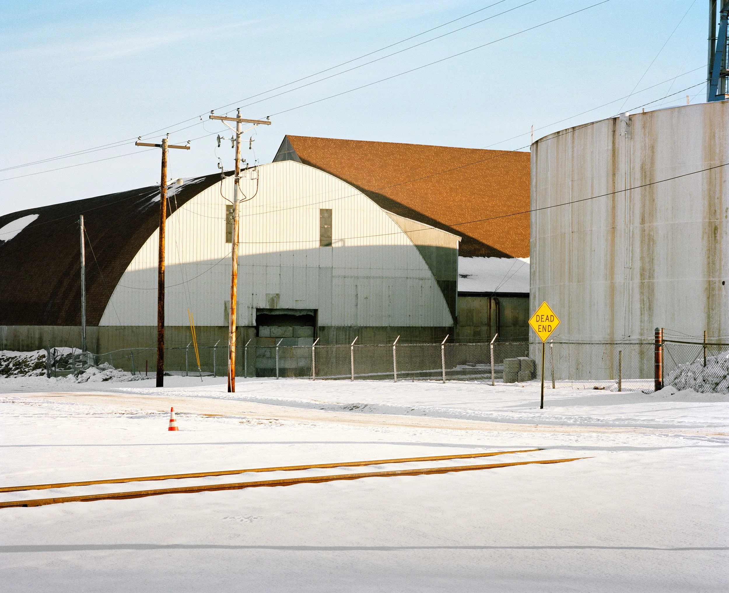 Industrial buildings and storage tanks behind a chain-link fence, with snow-covered ground, utility poles, and a yellow dead end sign, under a clear sky.