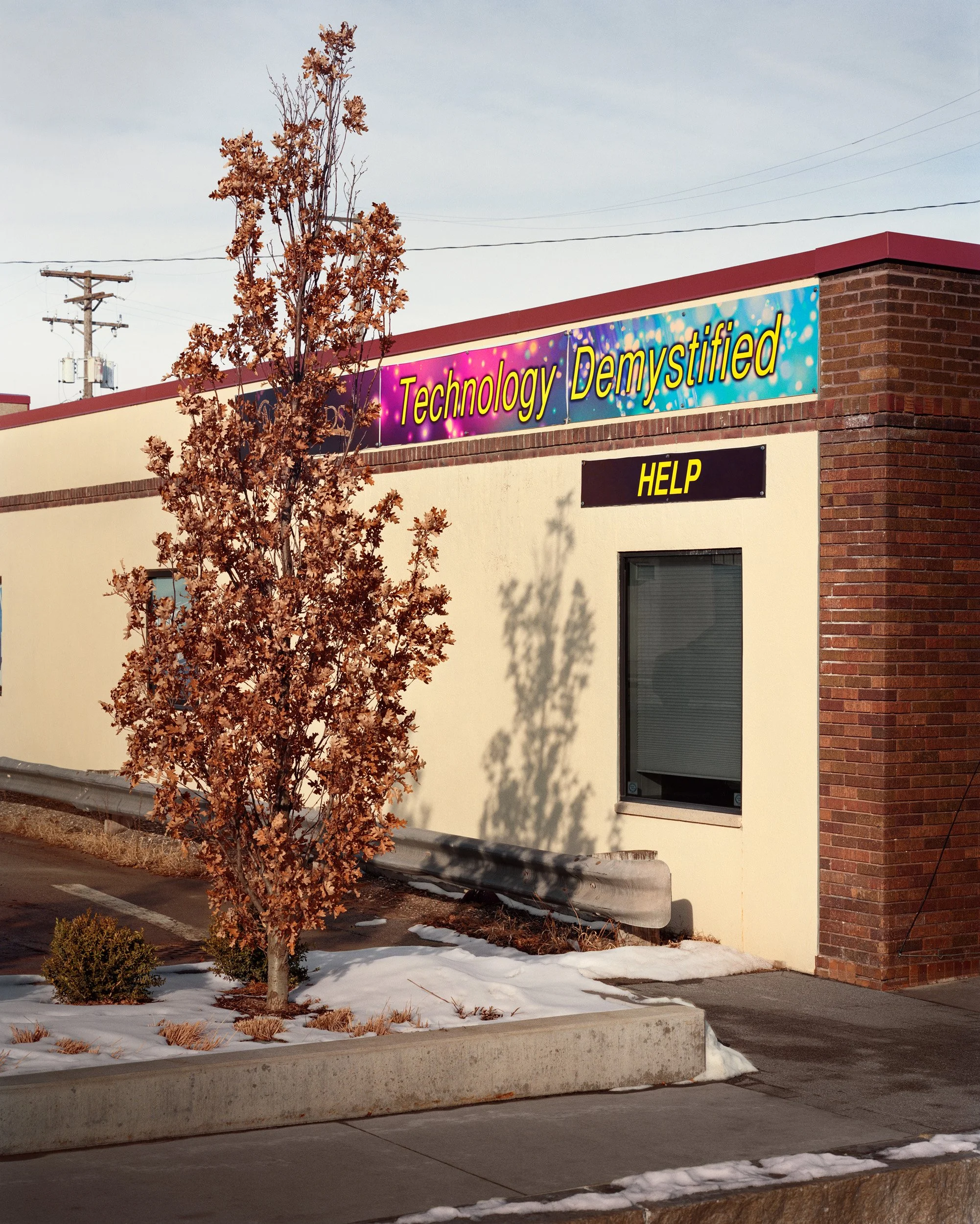 A building with a sign that reads 'Technology Demystified' and a black and yellow 'HELP' sign. There is a tree with brown leaves in front, snow on the ground, and power lines in the background.