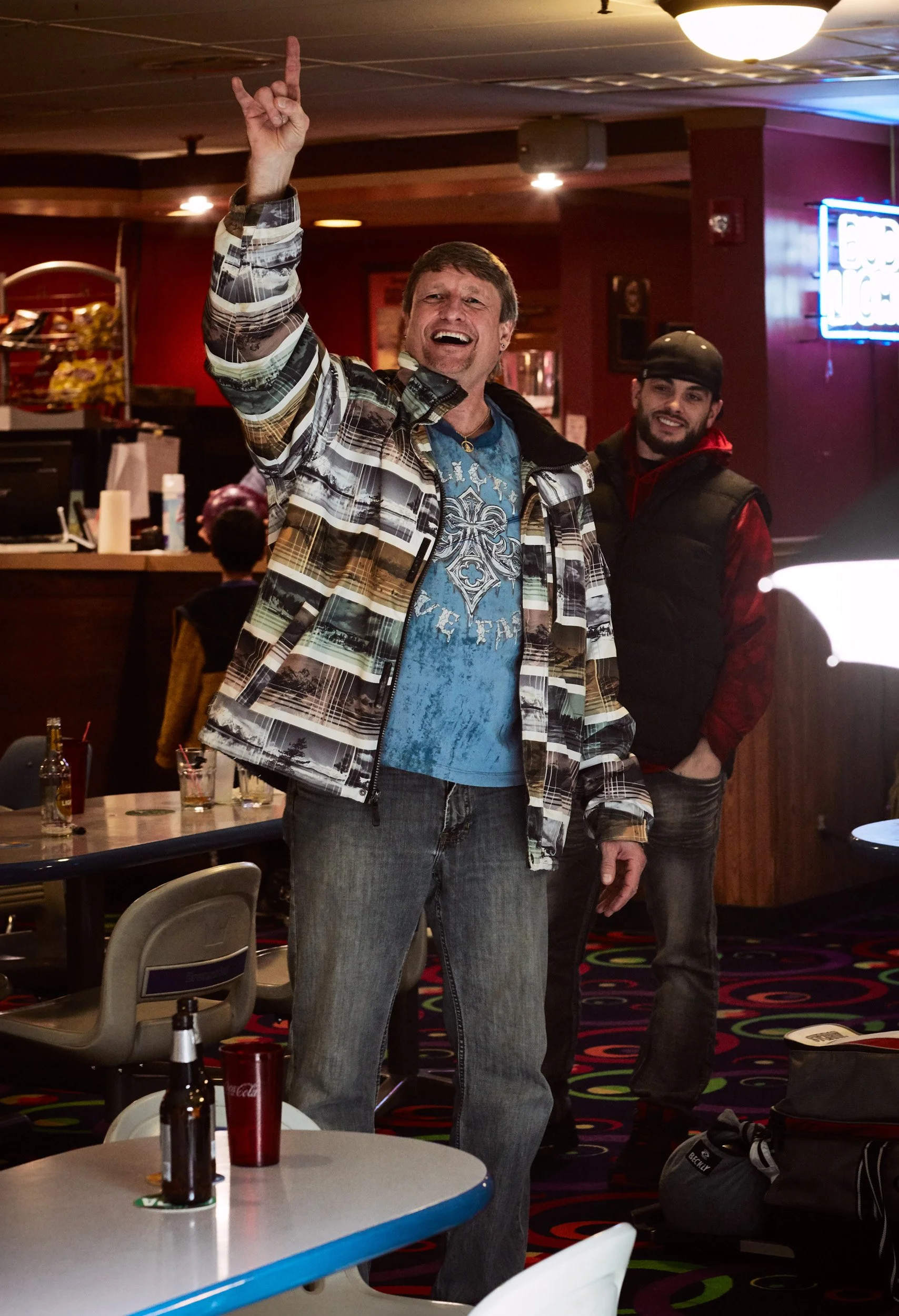 A man with short brown hair, wearing a patterned jacket and blue graphic T-shirt, is raising his right hand with his index finger pointing up and appears to be singing or cheering, standing in a lively bar or restaurant. There is another man in a bla