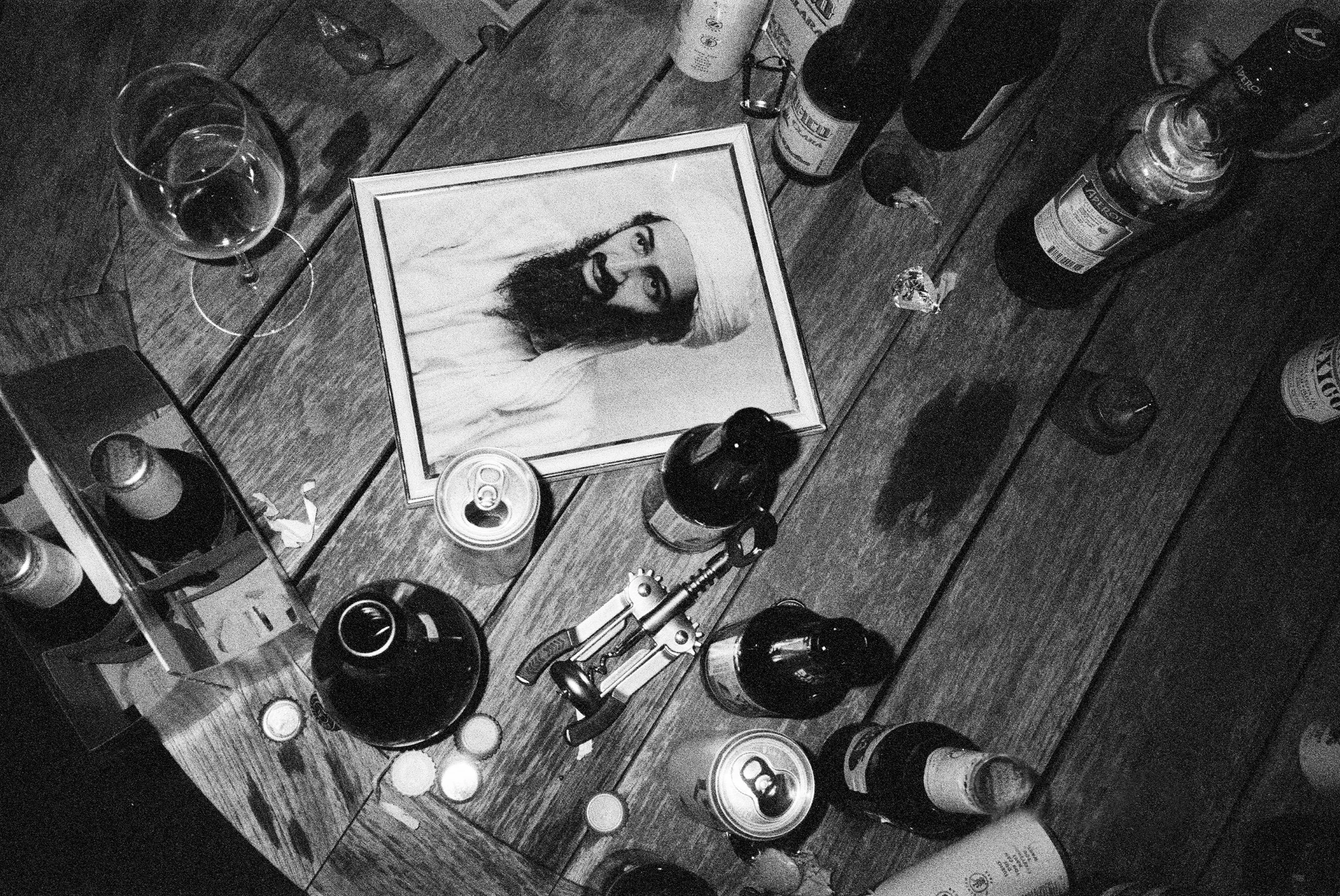 A black and white photo of a wooden table with various items, including a framed photo of a woman with long hair and a beard, several bottles, cans, and glasses, some candles, and a pair of keys.