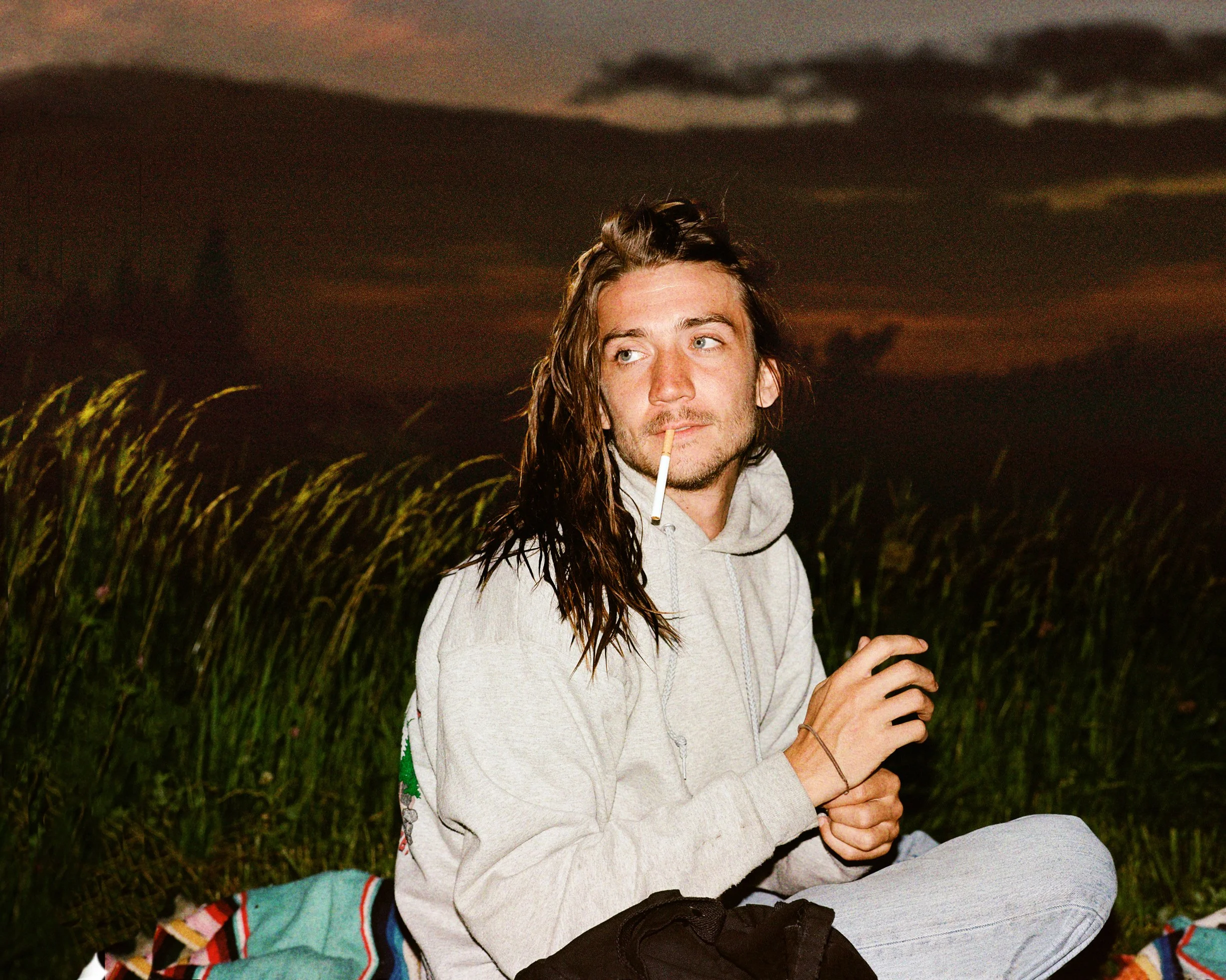 A young man with long hair and light facial hair sitting outdoors at sunset, wearing a gray hoodie and smoking a cigarette.