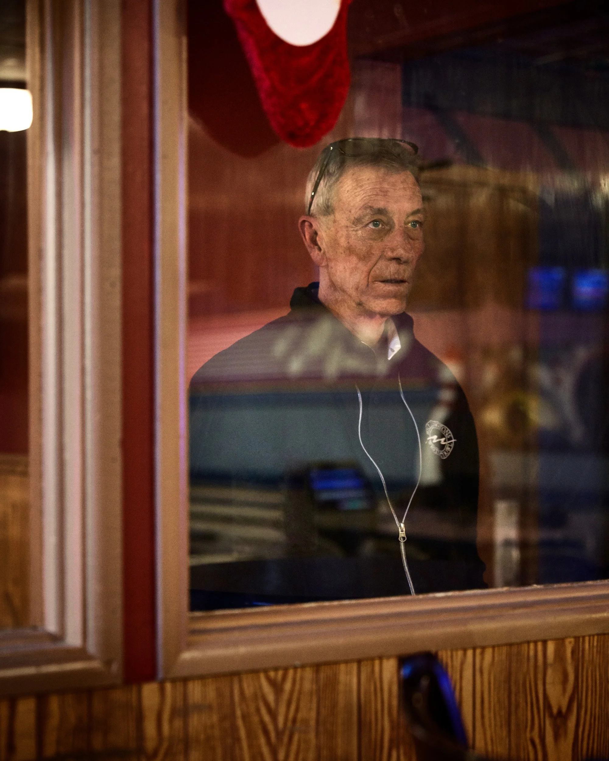 A man looking through a glass window at an indoor setting, with a red stocking hanging on the wall behind him.