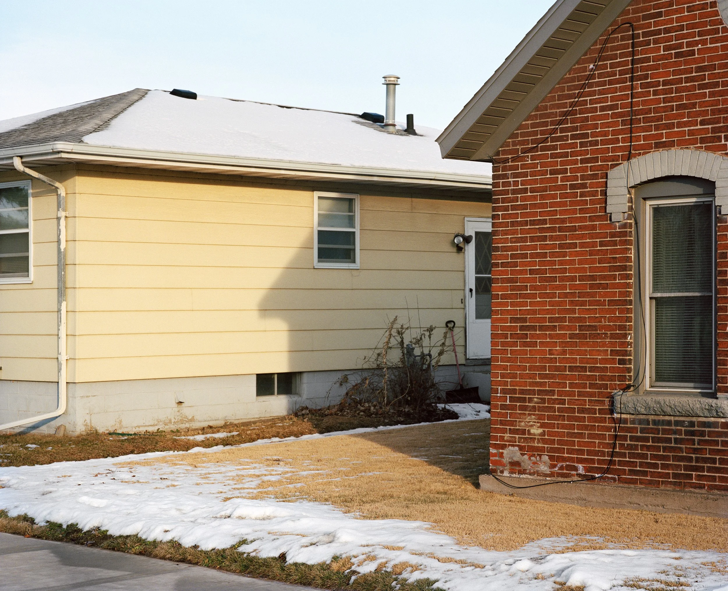 Side view of two houses, one yellow with siding, one red brick, with snow on the ground and a clear sky.