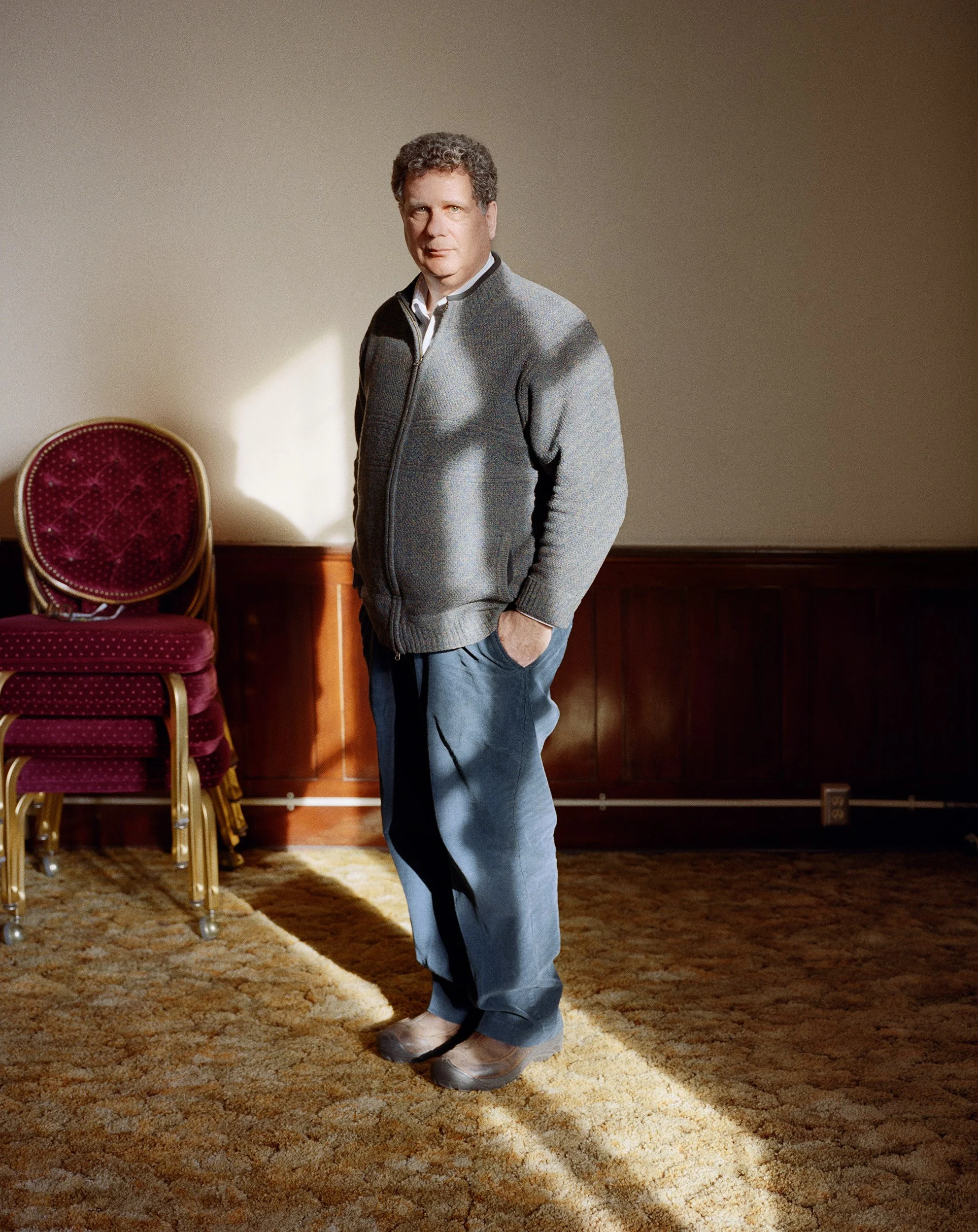 A man standing in a room with a stack of red chairs in the background, wearing a gray zip-up sweater, blue jeans, and brown shoes, with sunlight casting a shadow on the wall.