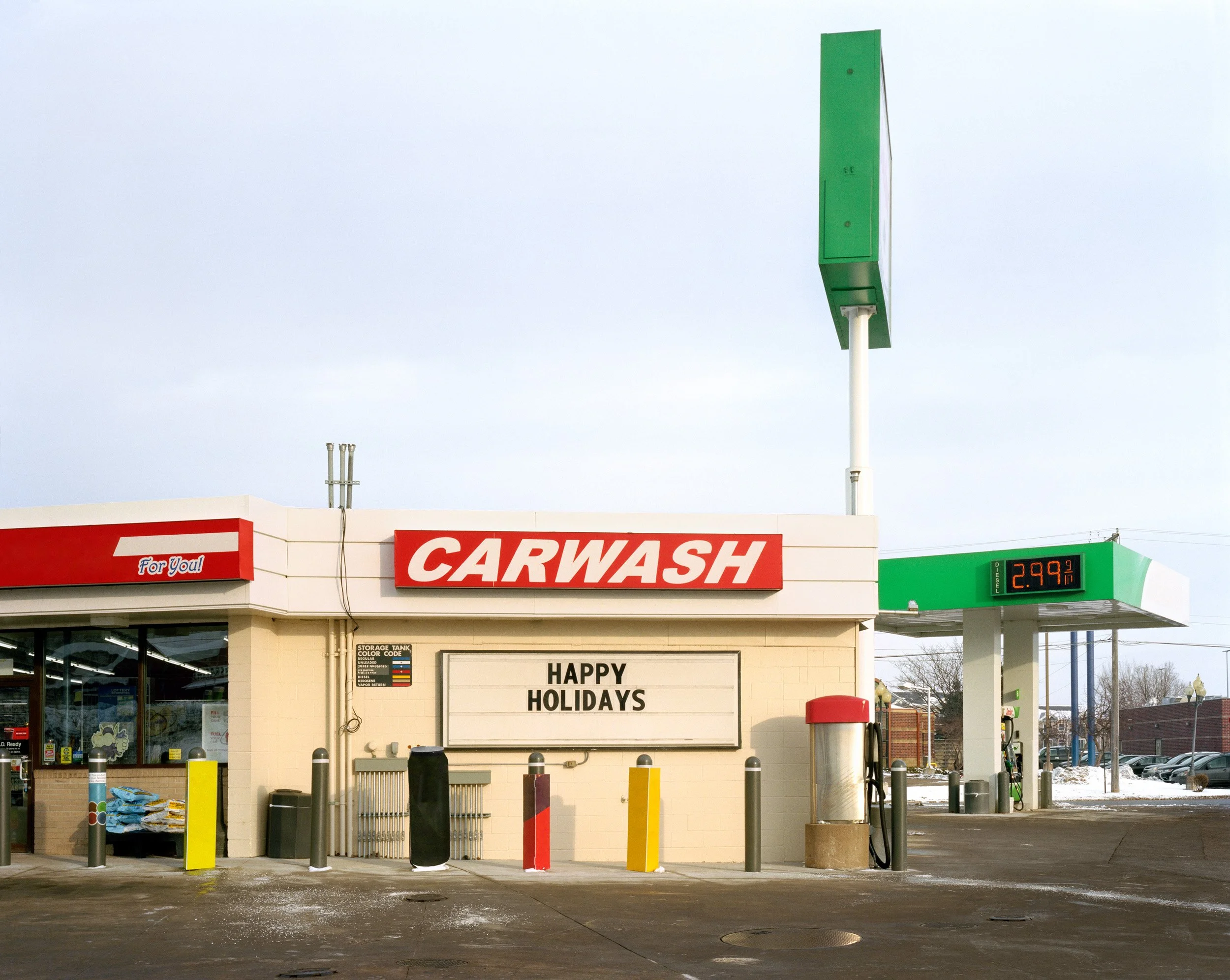 A car wash station with a sign saying "CARWASH" and a message board reading "HAPPY HOLIDAYS." There is a fuel price display showing 2.99 per gallon, a fueling pump, and a small store with a glass door and windows.