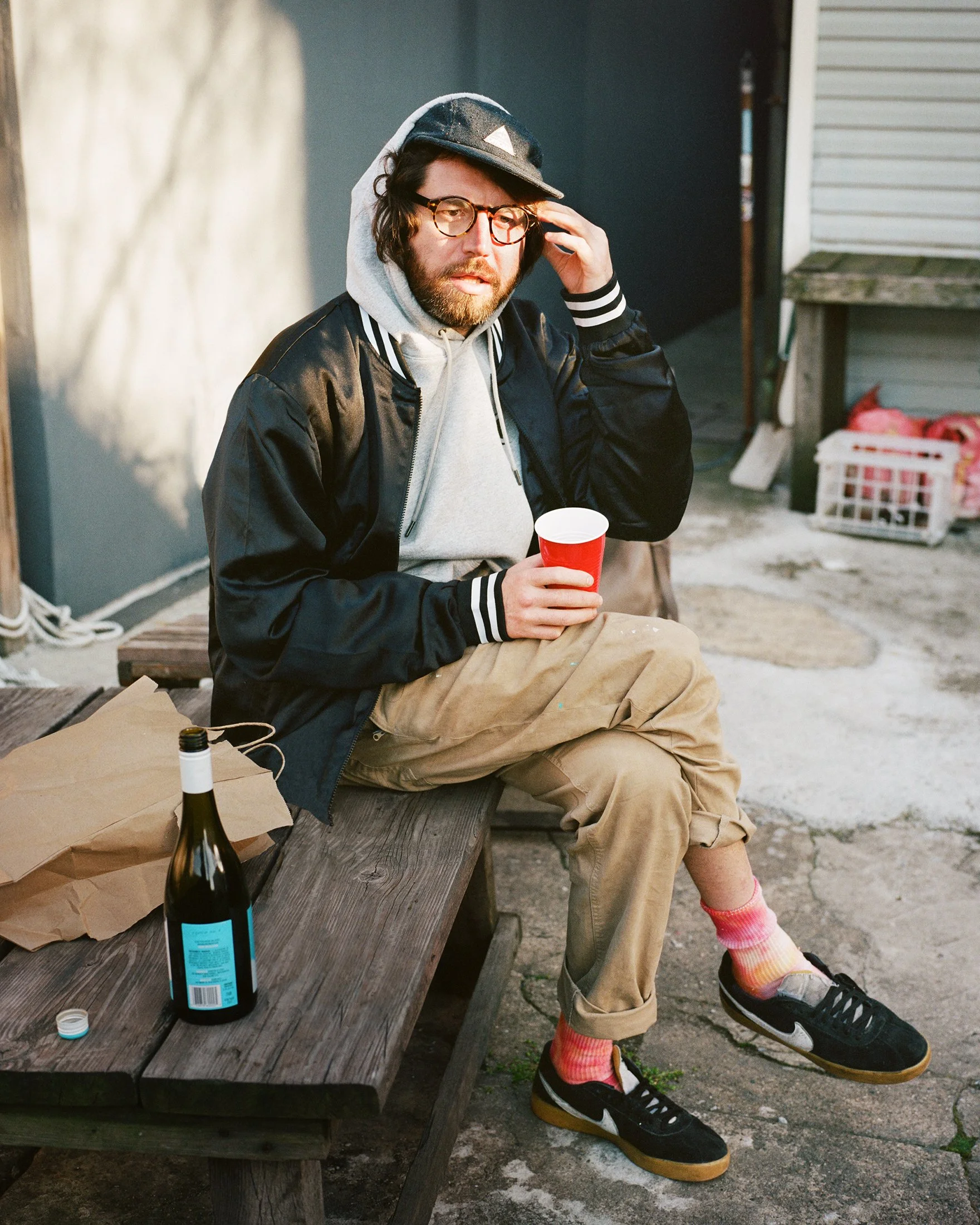 A man with glasses, beard, and long hair, dressed in a gray hoodie, black jacket, tan pants, and black sneakers with white soles, sits on a wooden bench outside, holding a red cup in his left hand and adjusting his glasses with his right hand. A bott