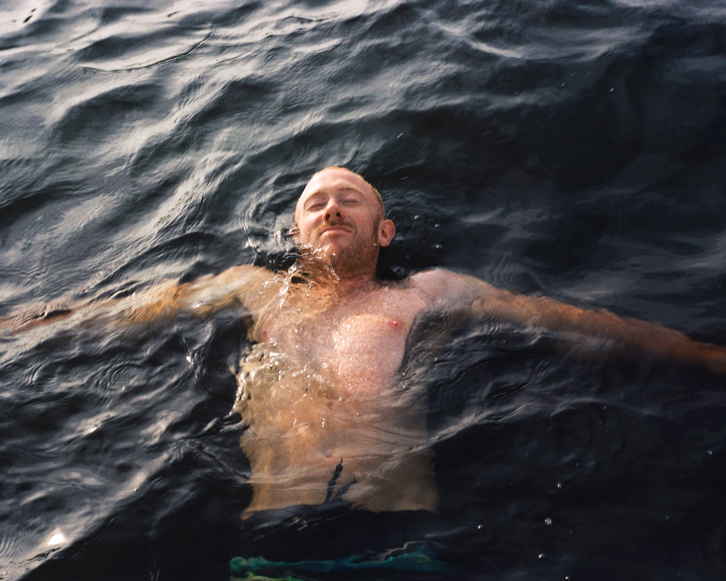 A man floating in dark water with his eyes closed and head tilted back.