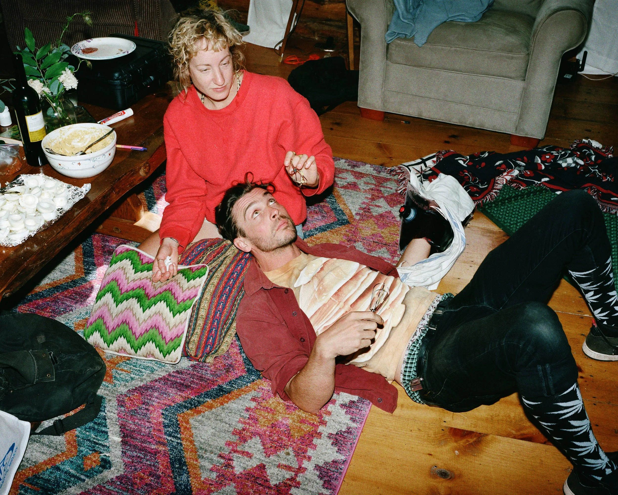 A woman in a red sweater kneeling next to a man lying on the floor, resting on pillows, with a colorful rug underneath. The man has dark hair, is wearing a red shirt, black jeans, and patterned tights. They are in a living room with a coffee table fi
